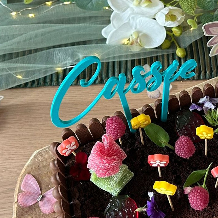 Gros plan sur le cake topper personnalisé modèle Élégance en bleu turquoise avec l'écriture "Cassie", piqué sur le dessus d'un gâteau d'anniversaire joliment décoré.