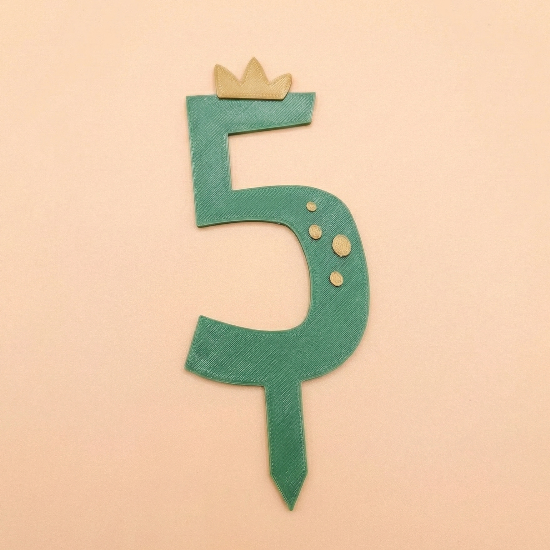 Decoration de gateau en forme de chiffre 5 vert avec une couronne miel et des taches en relief, presentee a plat sur un fond saumon.