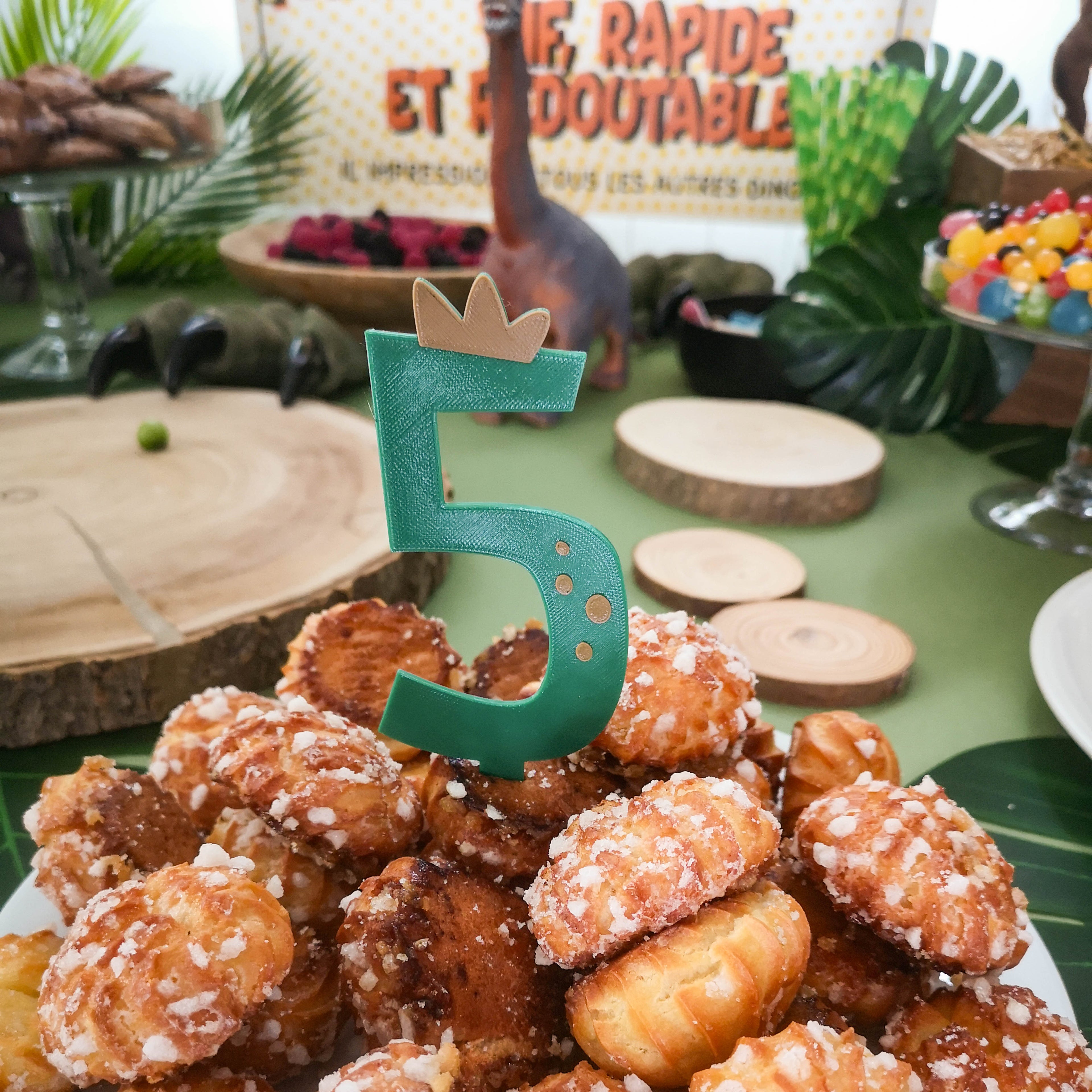 Pic de decoration pour gateau d anniversaire en forme de chiffre 5 vert avec couronne miel et reliefs de dinosaure plante dans un buffet de chouquettes theme jurassique.