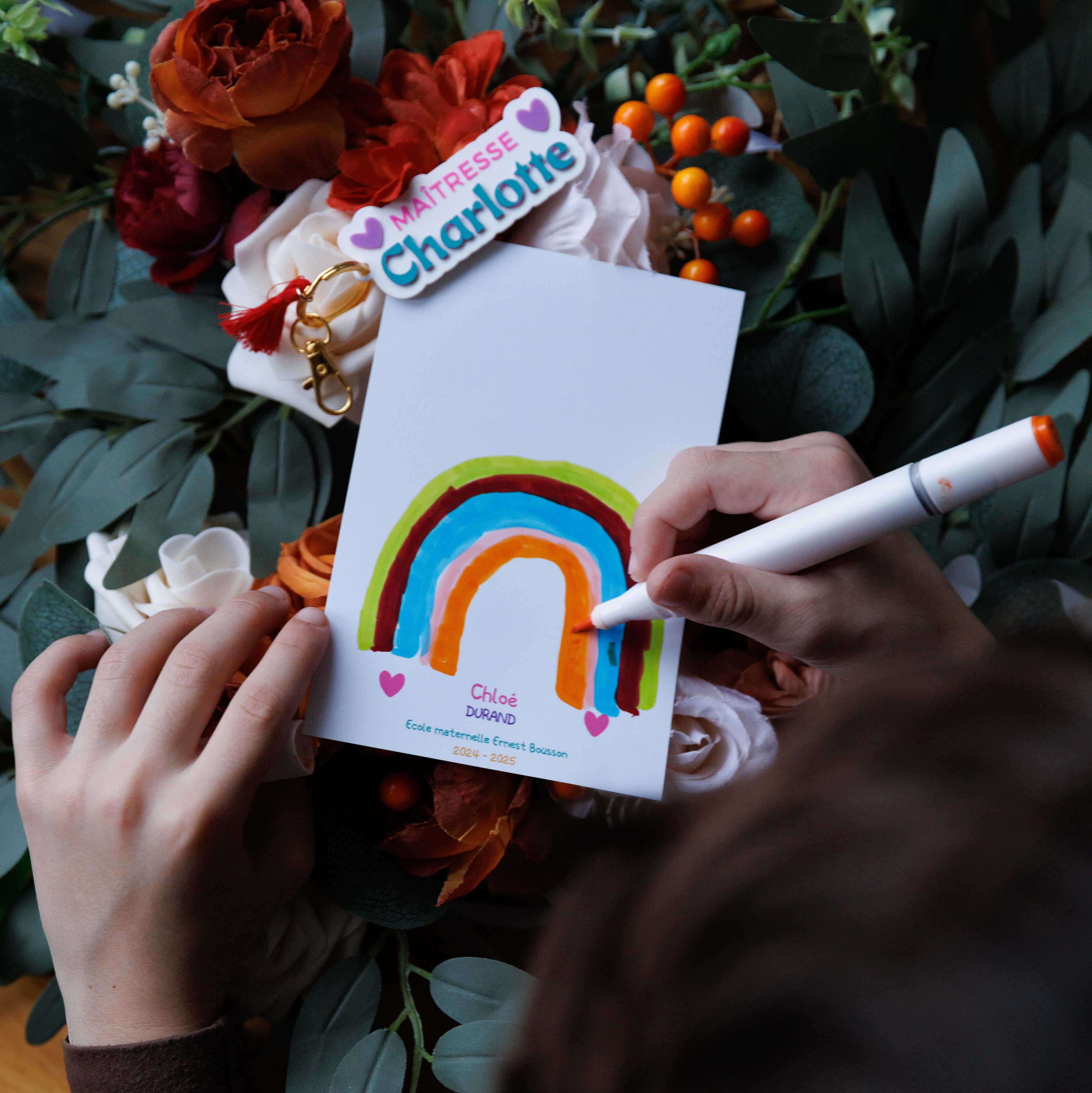 Photographie flat lay en haute définition montrant les mains de l'enfant Chloé Durand en train de colorier un arc-en-ciel sur une carte de remerciement blanche avec un marqueur orange. Sous le dessin de l'arc-en-ciel (vert, marron, bleu, rose), le texte imprimé est « Chloé », « DURAND », « Ecole maternelle Ernest Bousson » et « 2024-2025 ». Au-dessus de la carte, posée sur le bouquet, se trouve le porte-clef personnalisé.