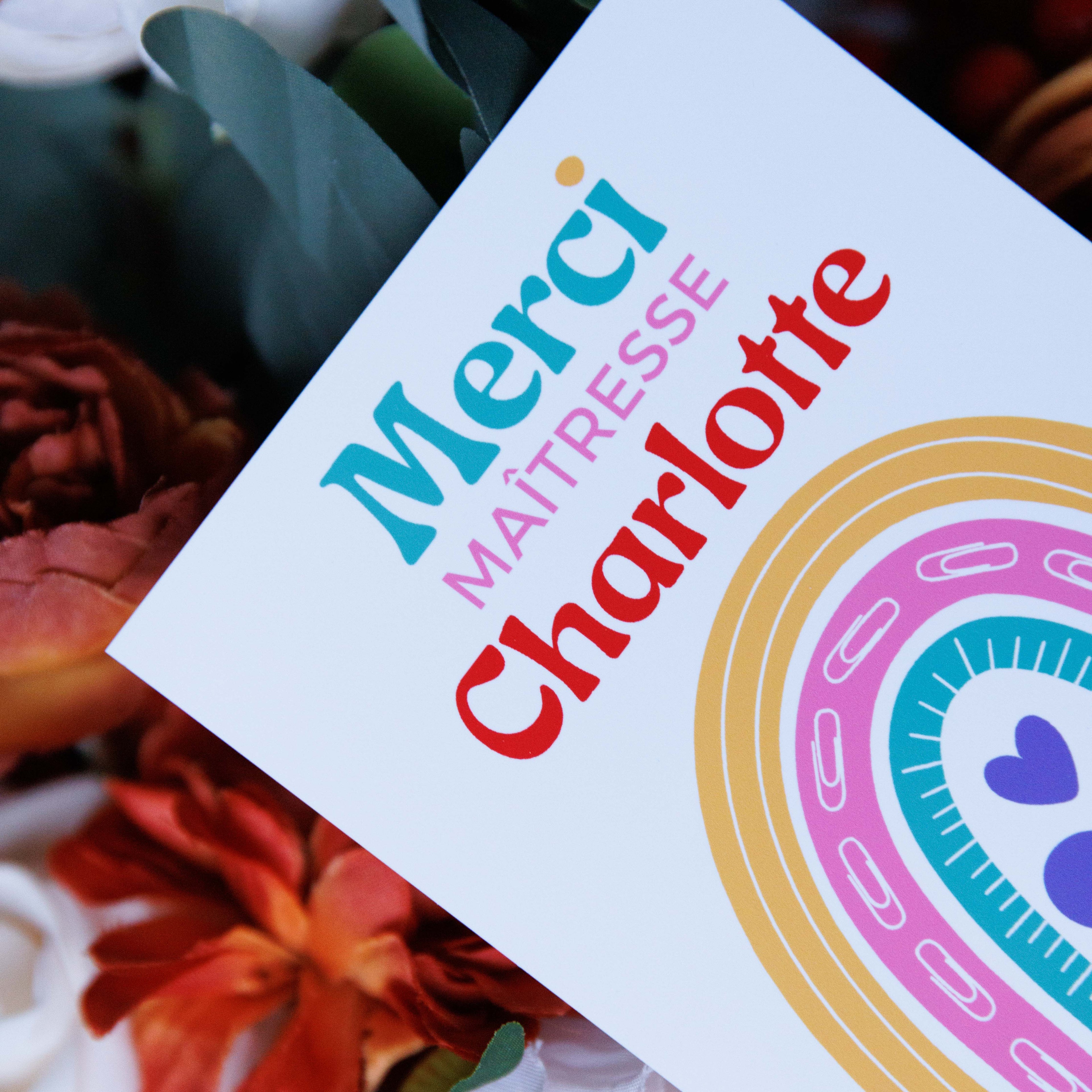 Gros plan d'une carte de remerciement personnalisée au nom de 'Charlotte', posée sur un bouquet de fleurs séchées de couleur rouille et de feuilles d'eucalyptus. La carte affiche le texte coloré : 'Merci' en bleu-vert, 'MAÎTRESSE' en rose, et 'Charlotte' en rouge. Un arc-en-ciel stylisé avec des trombones et des cœurs est visible.