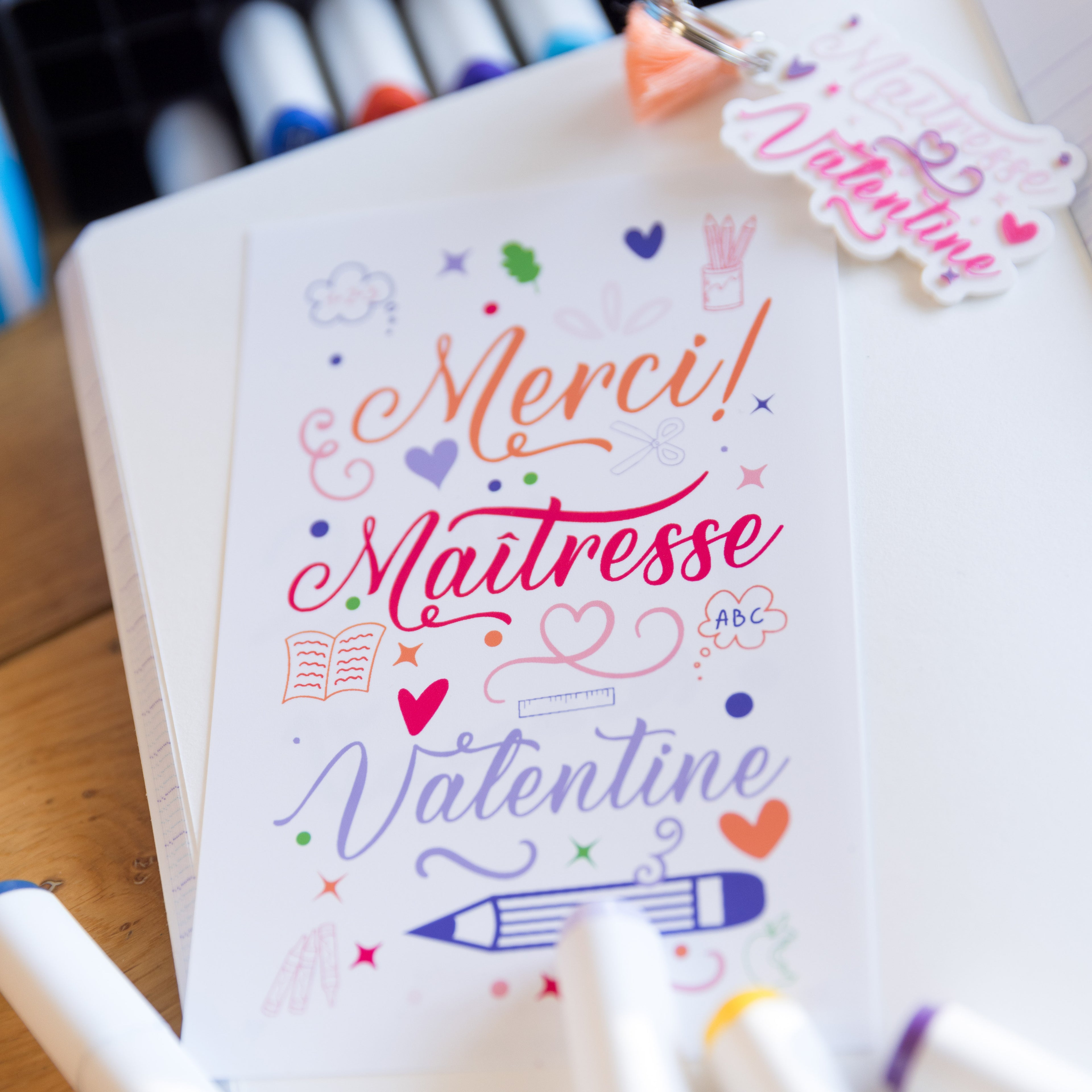 Carte de remerciement personnalisée avec le message « Merci ! Maîtresse Valentine » écrit dans des polices scriptes colorées. Le design « Savoirs et Arabesques » inclut des illustrations d'école comme un crayon bleu, un livre orange, une règle, des cœurs et des étoiles. La carte est posée sur un carnet blanc entourée de feutres de dessin.