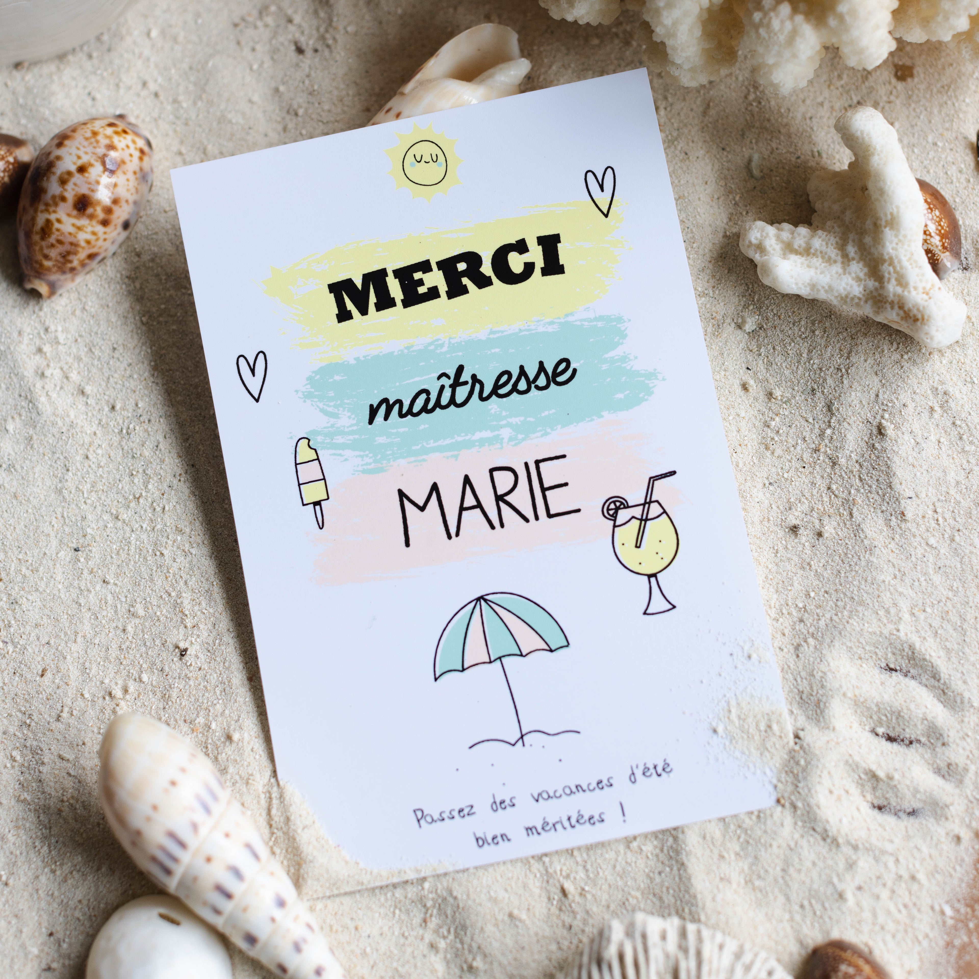 Gros plan d'une carte de remerciement personnalisée "Merci maîtresse Marie" sur le thème des vacances d'été. La carte est posée sur du sable fin entourée de coquillages et de coraux. Elle comporte le message "Passez des vacances d'été bien méritées !" ainsi que des illustrations de parasol, cocktail et glace sur des touches de couleurs pastel.