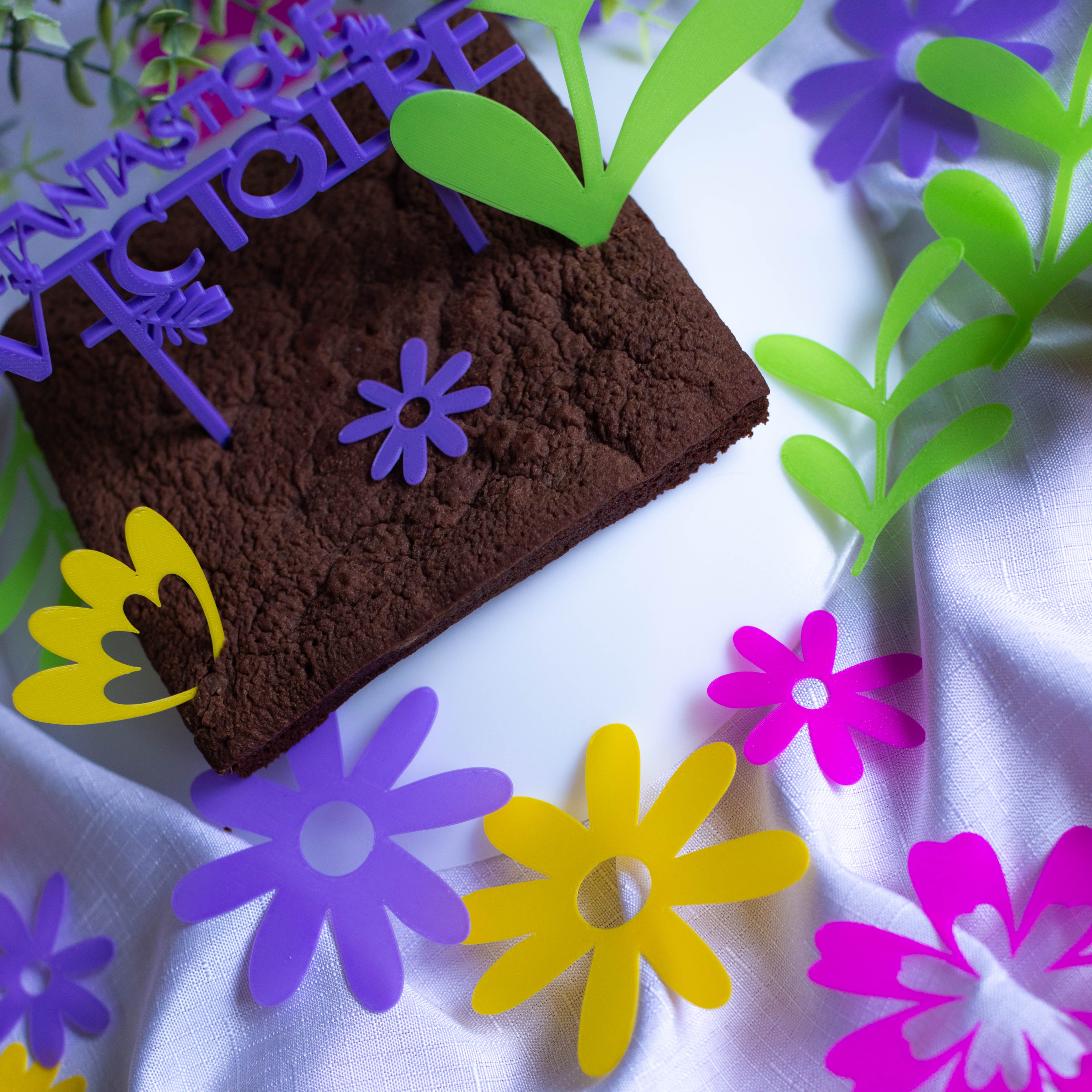 Mise en scène d'un gâteau au chocolat décoré de cake toppers et entouré de confettis de table Magie Florale en impression 3D PLA. Des fleurs et feuillages stylisés de couleurs jaune, violet, rose et vert pomme sont disposés sur une nappe blanche drapée autour du dessert.
