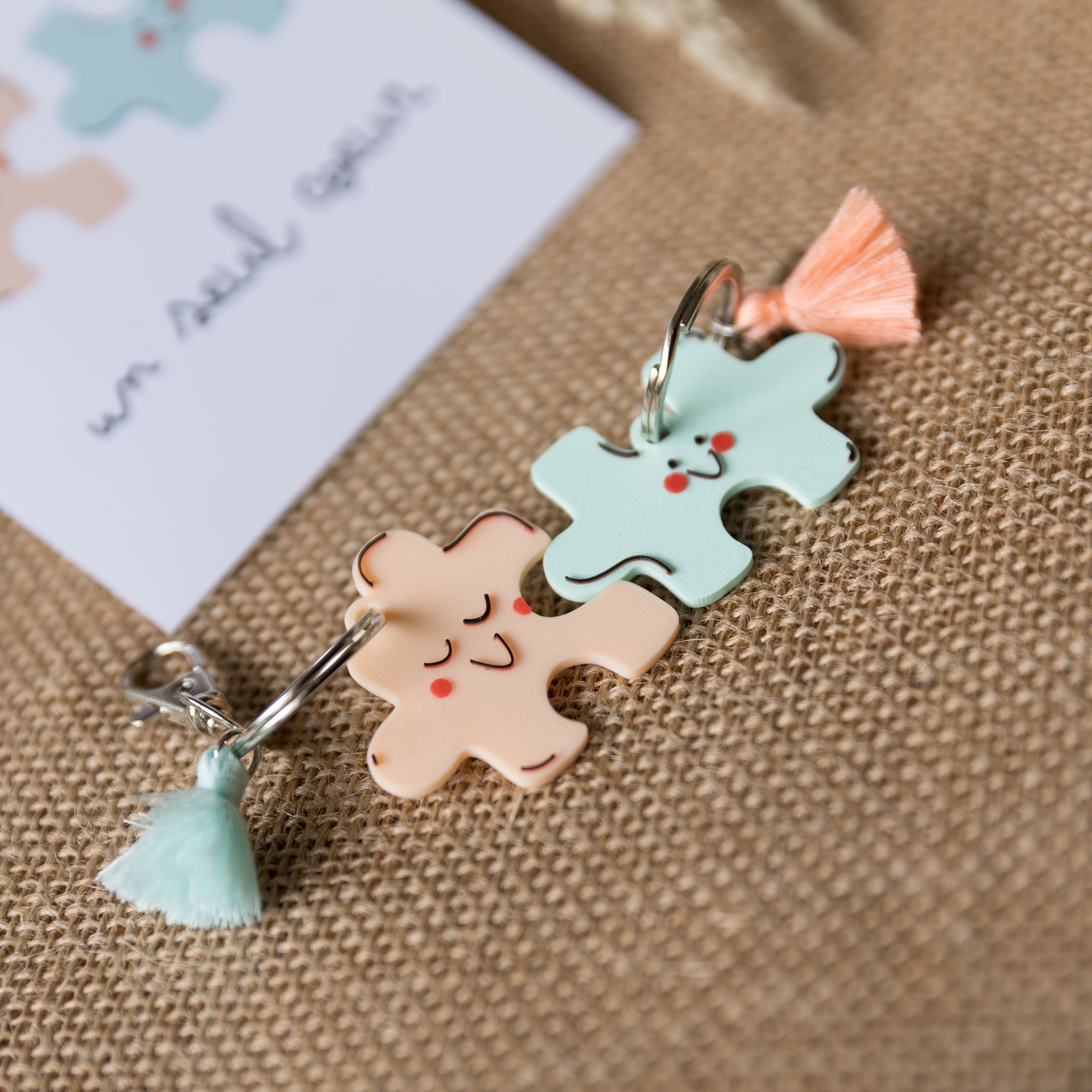 Gros plan de deux porte-clés en forme de pièces de puzzle kawaii, l'un de couleur pêche et l'autre menthe à l'eau, avec des petits pompons assortis. Les porte-clés sont disposés sur un fond de jute texturé avec une carte "un seul cœur" floue en arrière-plan.