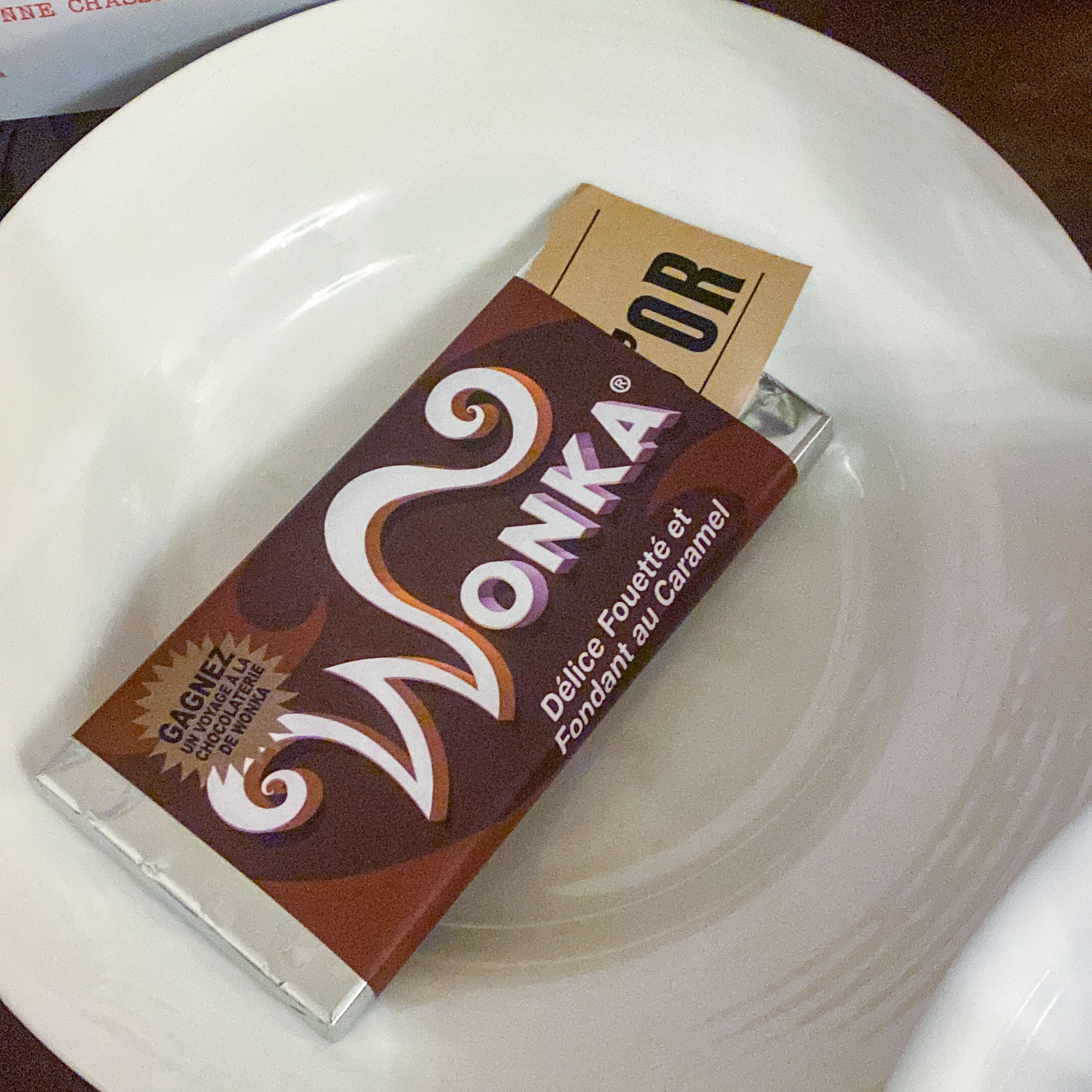 Gros plan sur une tablette de chocolat avec un emballage personnalisé "Wonka" et un "Ticket d'Or" dépassant de l'étui, le tout posé sur une assiette blanche. Une mise en scène réaliste issue du kit PDF pour recréer l'ambiance de Charlie et la chocolaterie.