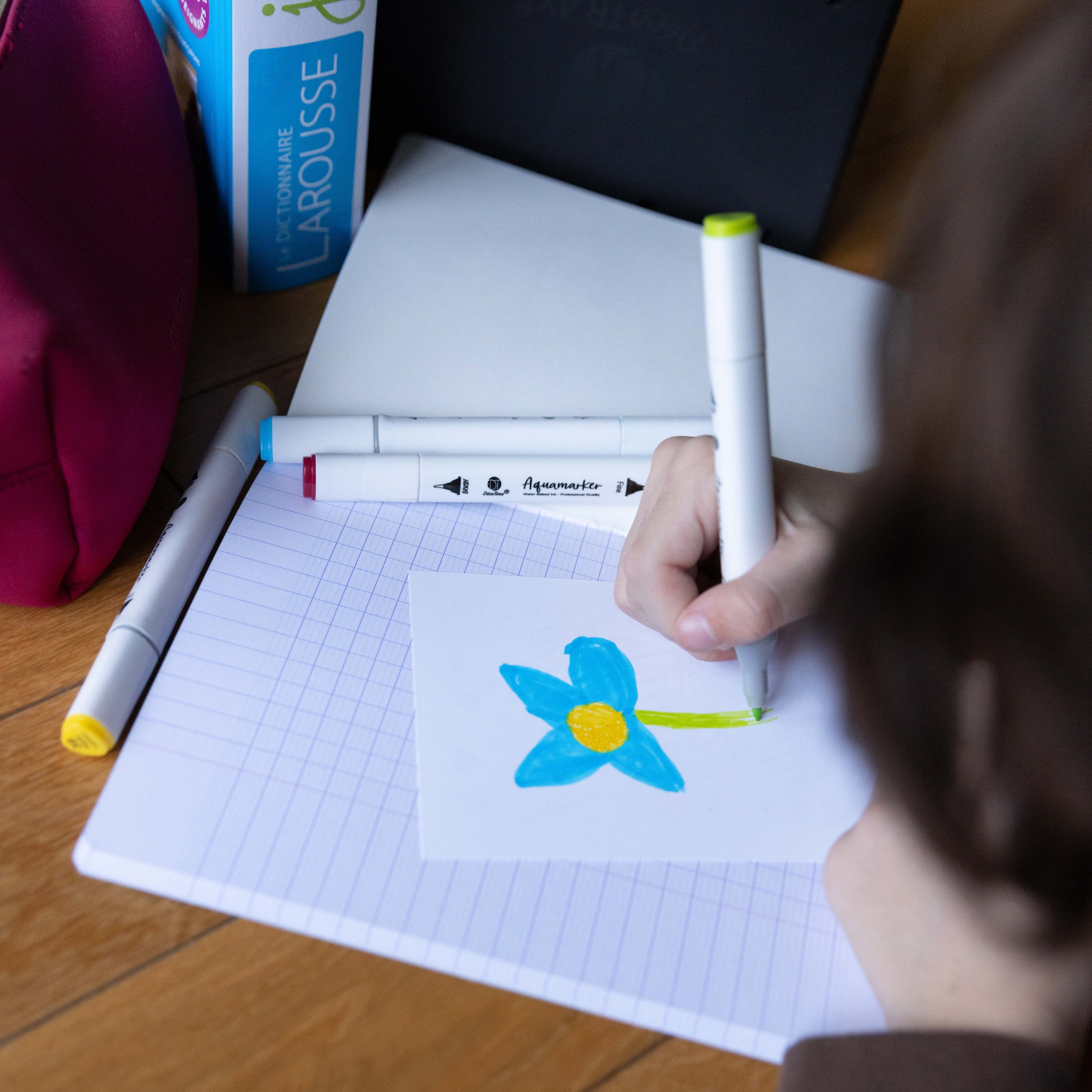 Vue de dessus d'un enfant en train de colorier une fleur bleue et jaune avec un feutre vert sur une carte de remerciement. La scène se passe sur un bureau avec un cahier à petits carreaux, des marqueurs, une trousse rose et un dictionnaire en arrière-plan.