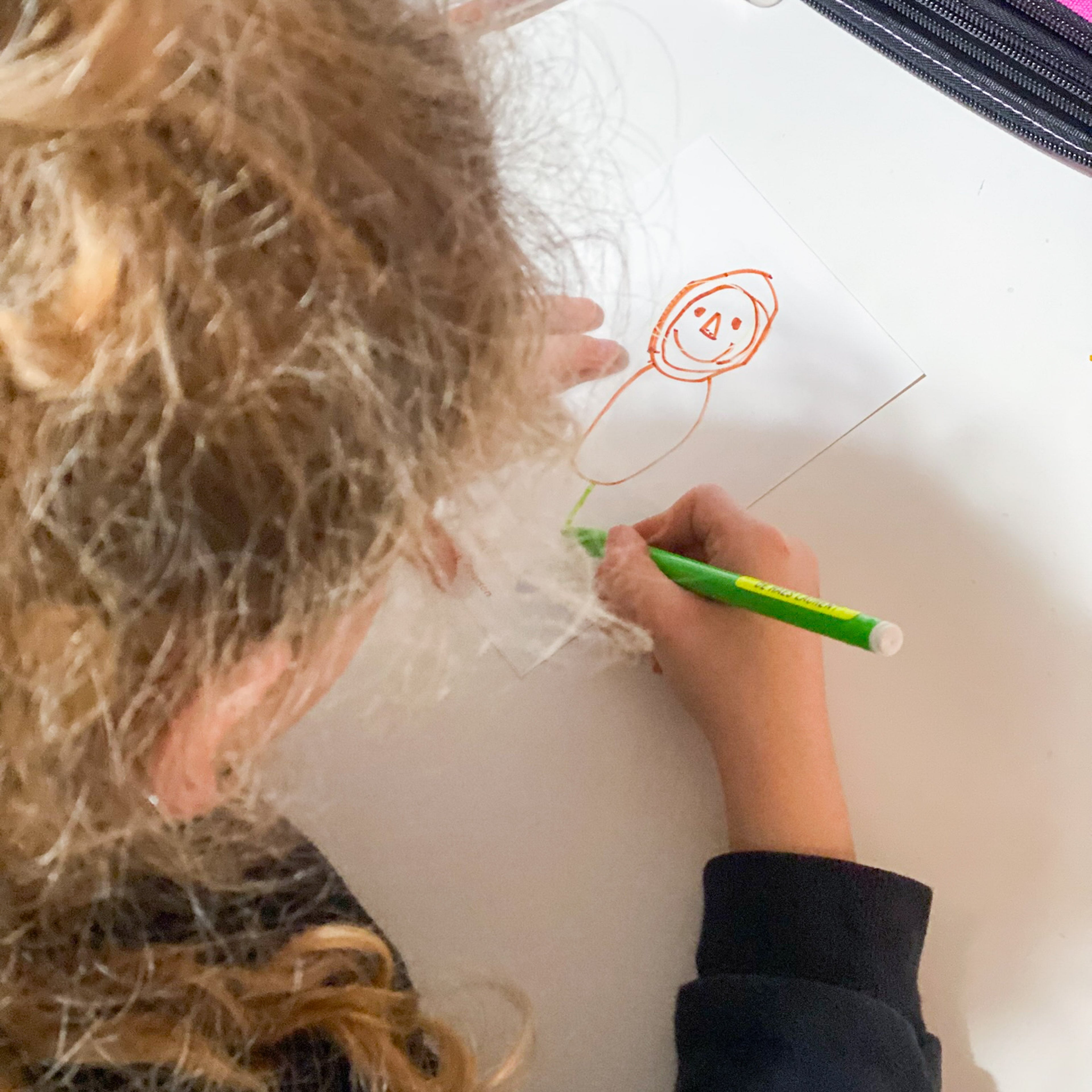 Vue de dessus d'un enfant aux cheveux bouclés en train de dessiner au verso d'une carte de remerciement blanche. Il utilise un feutre vert pour compléter un dessin de personnage tracé à l'orange. Cette photo montre l'étape de personnalisation créative du kit cadeau par l'enfant.