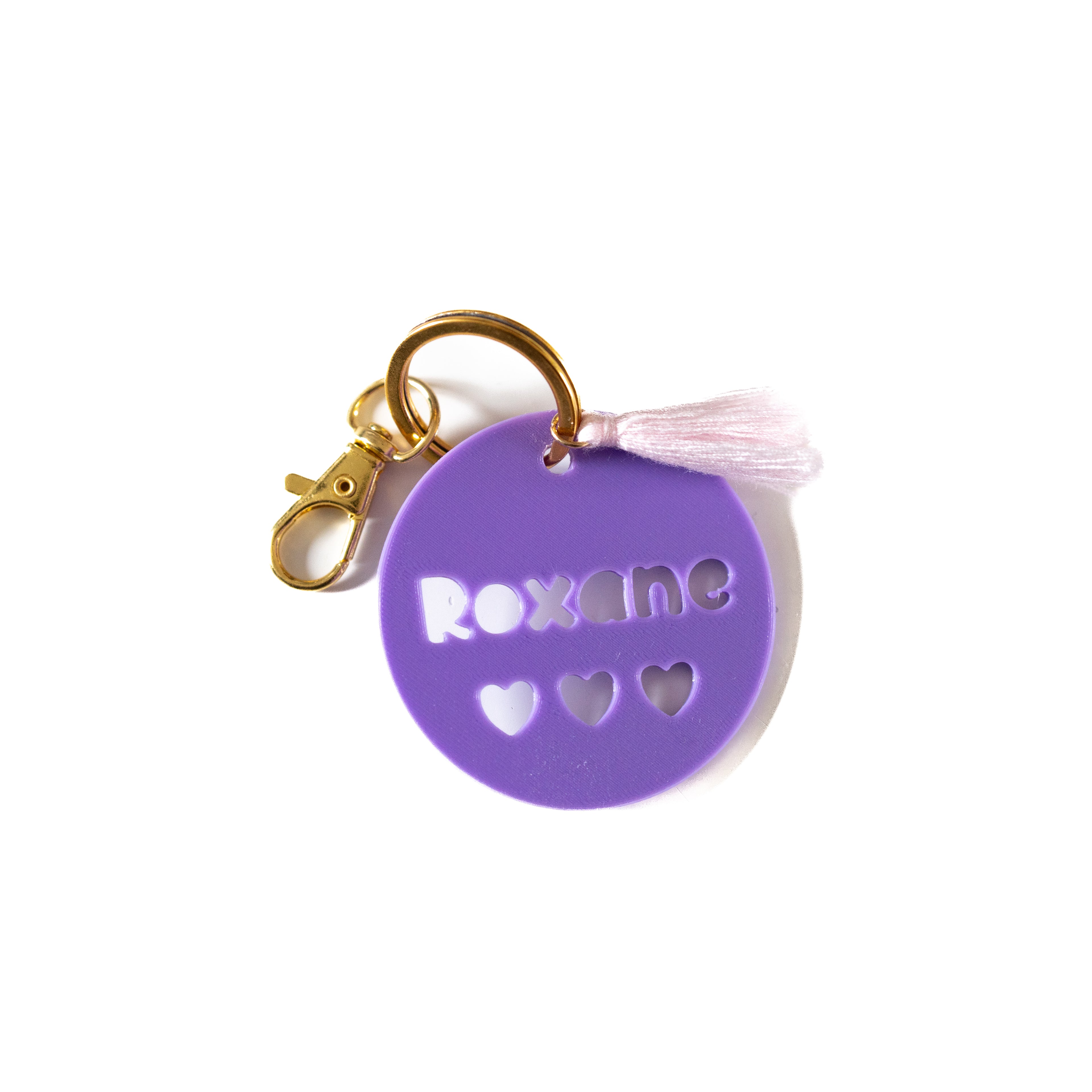 Vue premium d'une étiquette de sac 3D personnalisée en PLA violet, avec le prénom 'Roxane' découpé, un mousqueton doré et un pompon rose pastel sur fond blanc.