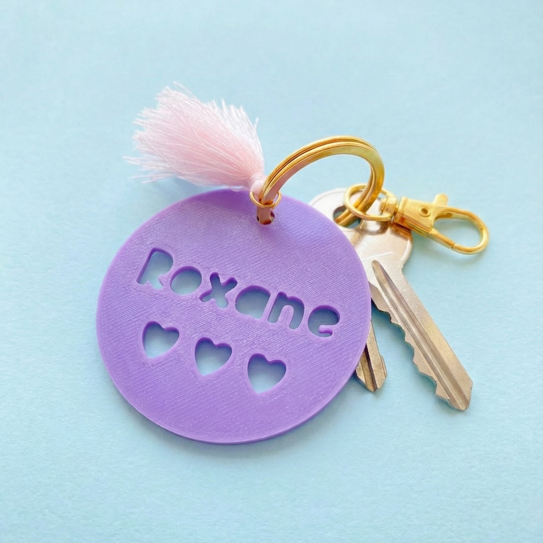 Gros plan sur un médaillon violet imprimé en 3D avec le prénom Roxane découpé, agrémenté d'un pompon soyeux rose et d'une attache en métal doré brillant.