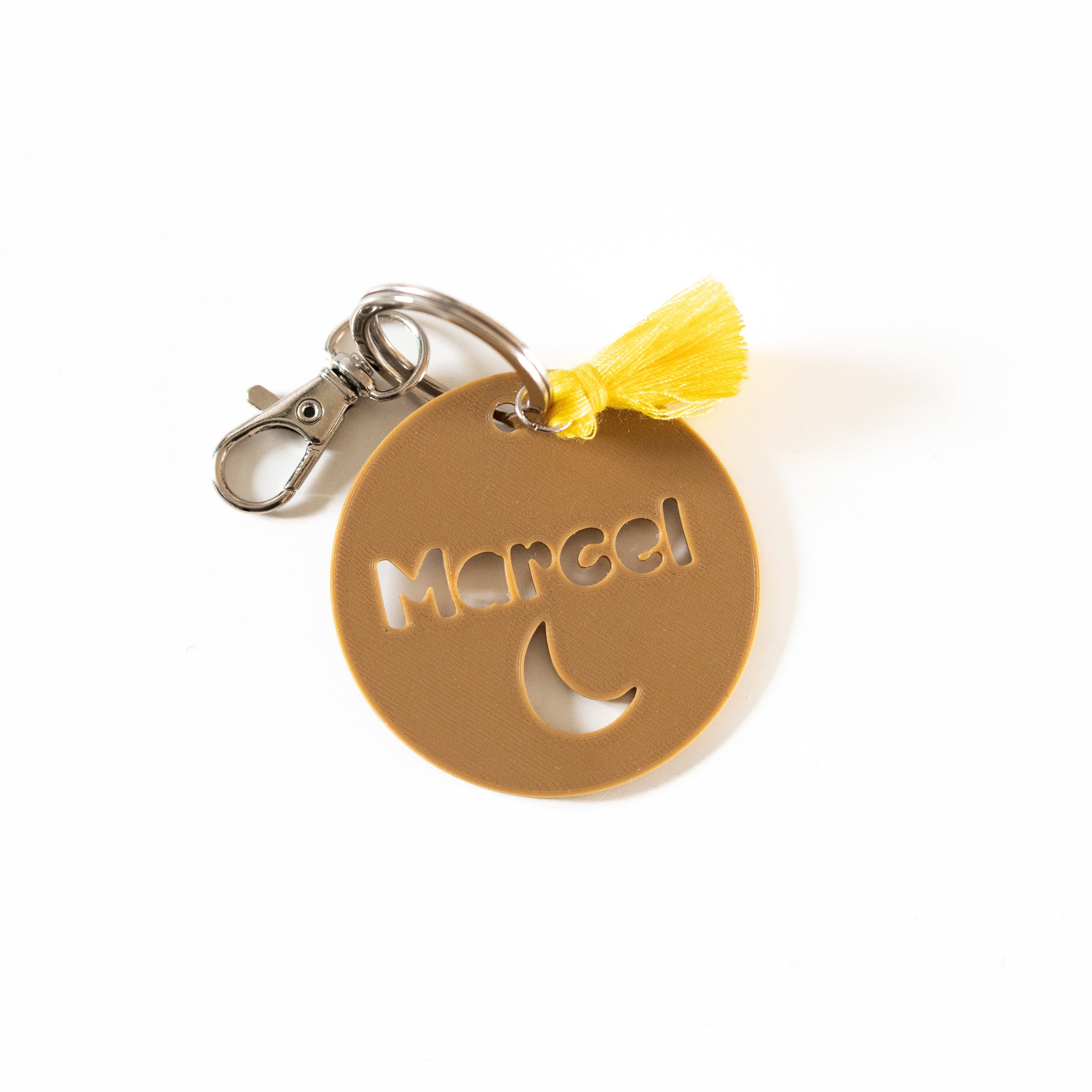 Vue rapprochée d'un médaillon personnalisé en impression 3D couleur café avec le prénom Marcel et un motif lune, accompagné d'un pompon jaune vif et d'un mousqueton argenté sur fond blanc.