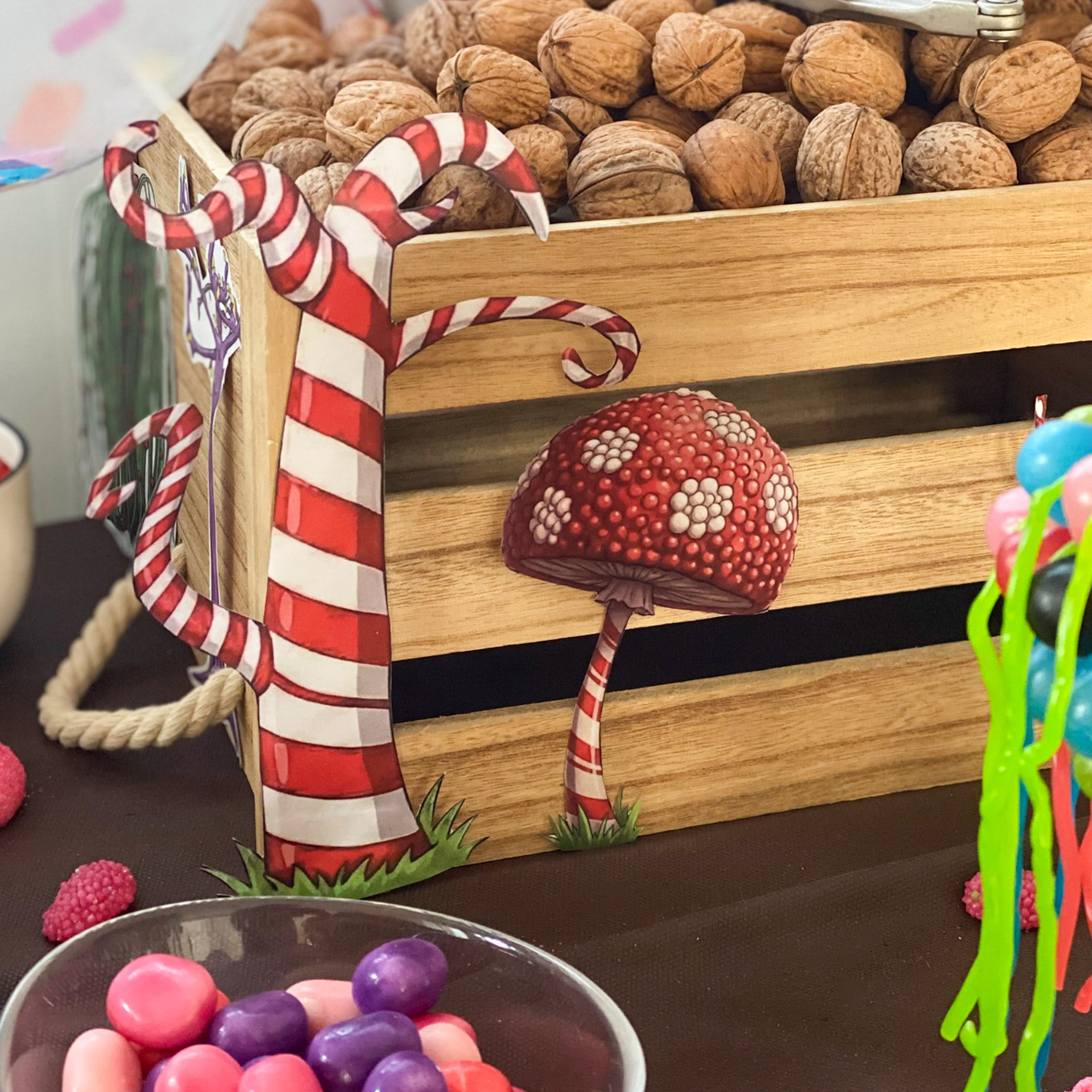 Gros plan sur une caisse en bois remplie de noix, décorée avec des illustrations découpées d'un arbre fantastique et d'un champignon à rayures rouges et blanches. Un bol de bonbons colorés au premier plan complète cette mise en scène créative issue d'un kit de décoration à imprimer.