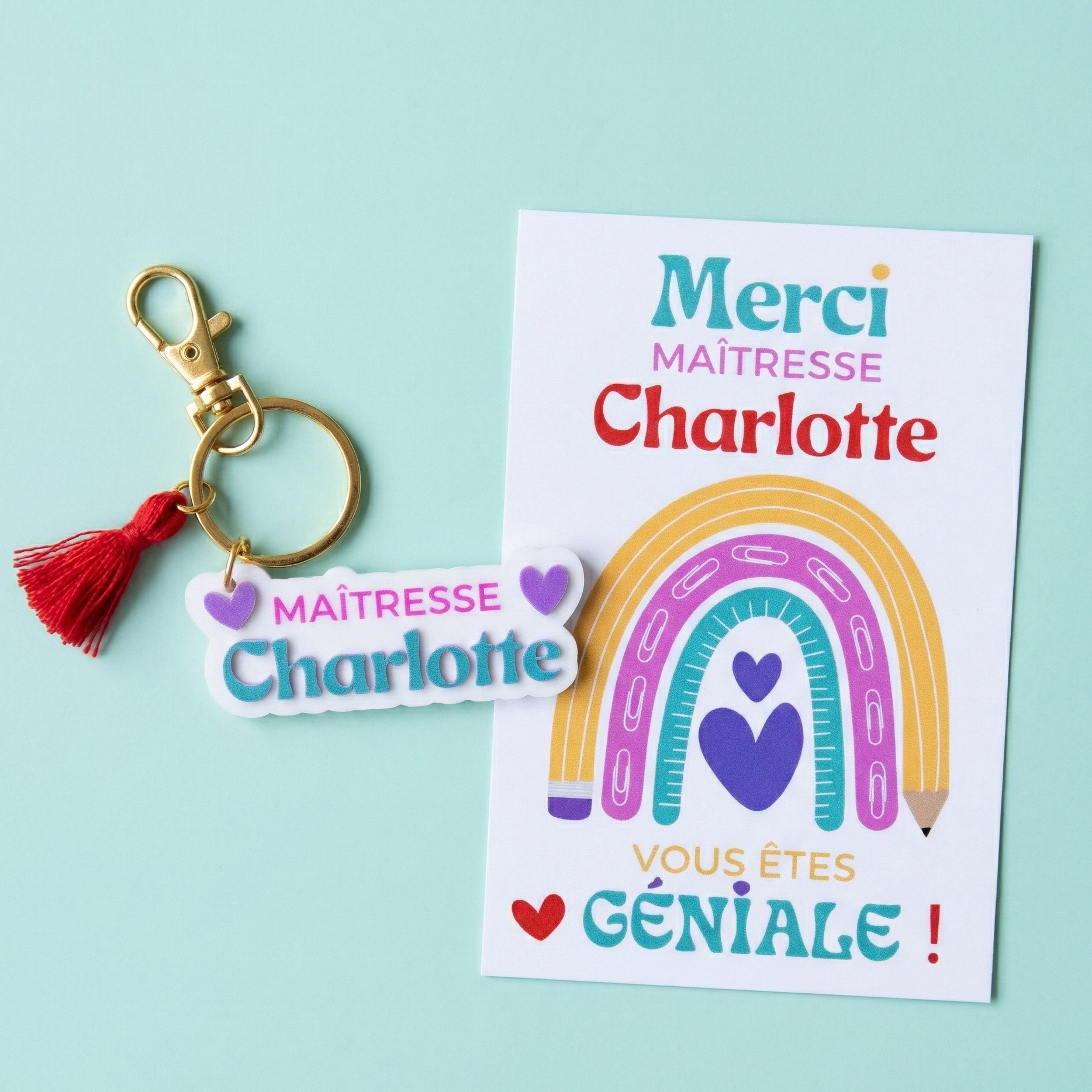 Kit cadeau personnalisé pour maîtresse posé sur un fond vert menthe, comprenant un porte-clés blanc avec texte 'Maîtresse Charlotte' et pompon rouge, accompagné d'une carte de remerciement assortie avec un arc-en-ciel coloré et le message 'Merci Maîtresse Charlotte, vous êtes géniale !'.