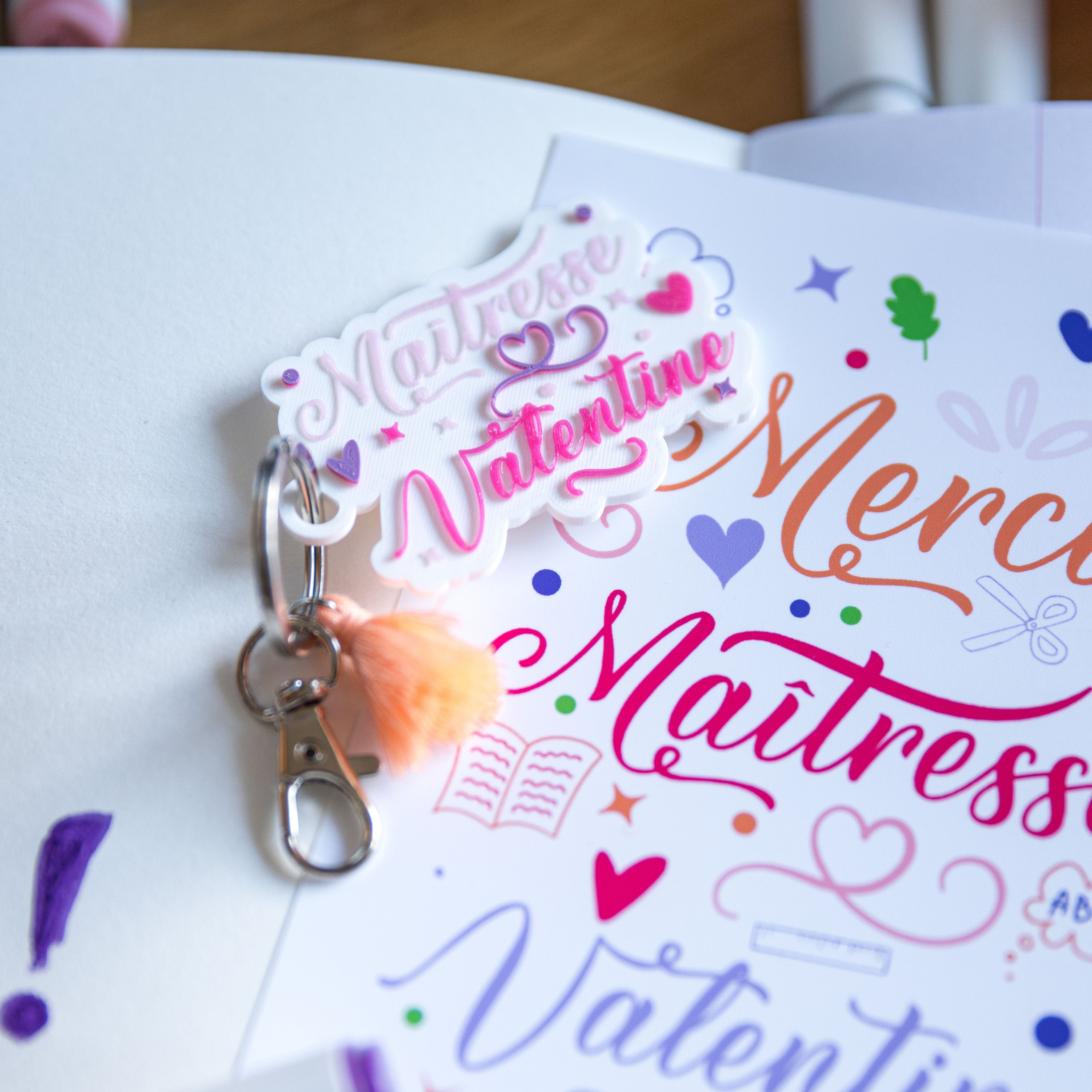 Gros plan d'un porte-clés personnalisé "Maîtresse Valentine" réalisé en impression 3D PLA blanc. Le texte apparaît en relief avec des teintes de rose et de violet, agrémenté de petits cœurs et d'étoiles colorées. L'attache argentée est munie d'un mousqueton et d'un pompon de couleur pêche, le tout posé sur une carte blanche.