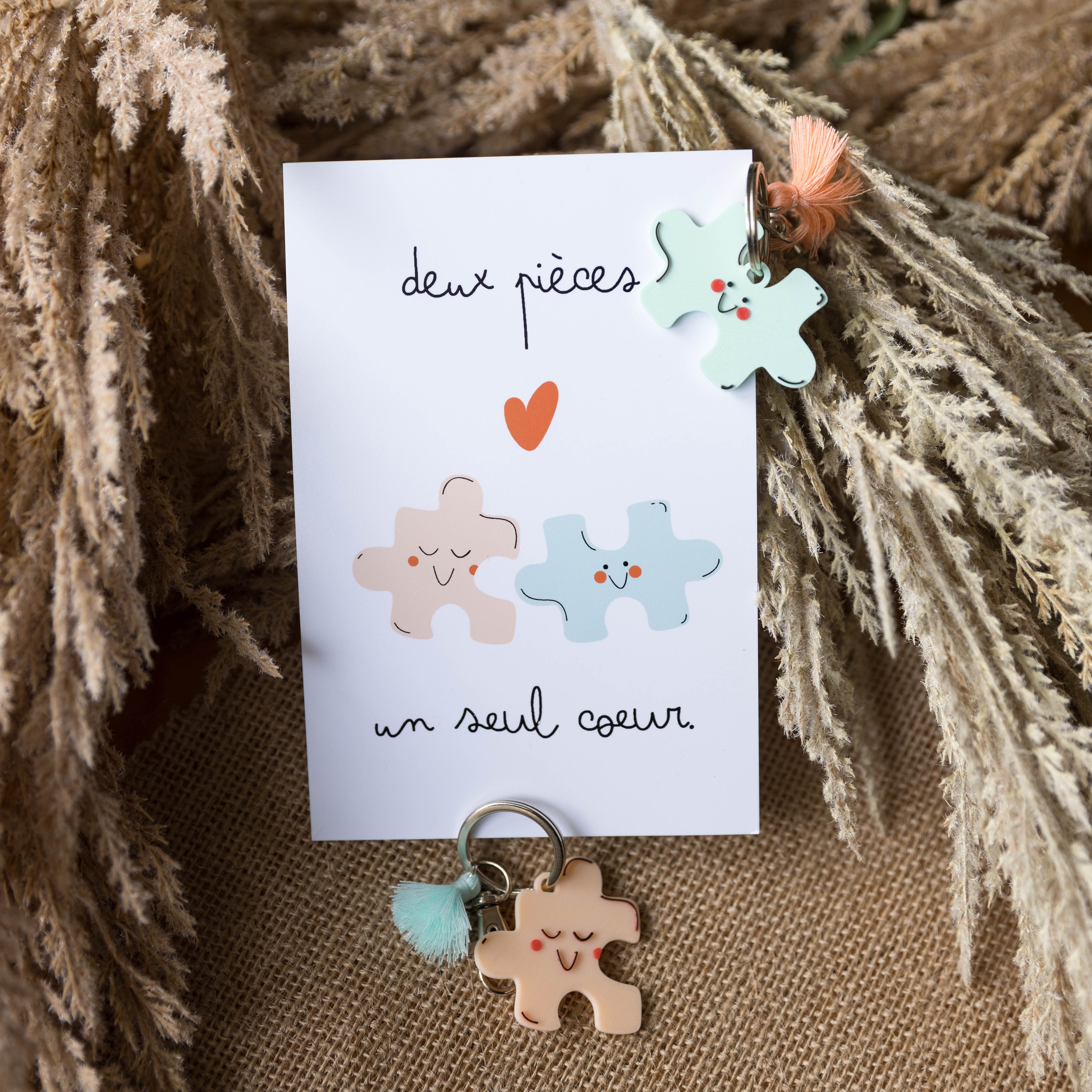 Kit cadeau duo composé d'une carte manuscrite 'deux pièces un seul cœur' et de deux porte-clés assortis en forme de pièces de puzzle avec des visages souriants (kawaii) et des petits pompons (un menthe avec pompon rose, un rose pêche avec pompon menthe). Le kit repose sur un fond de toile de jute et d'herbe de pampa séchée, pour un cadeau de couple bohème et mignon.