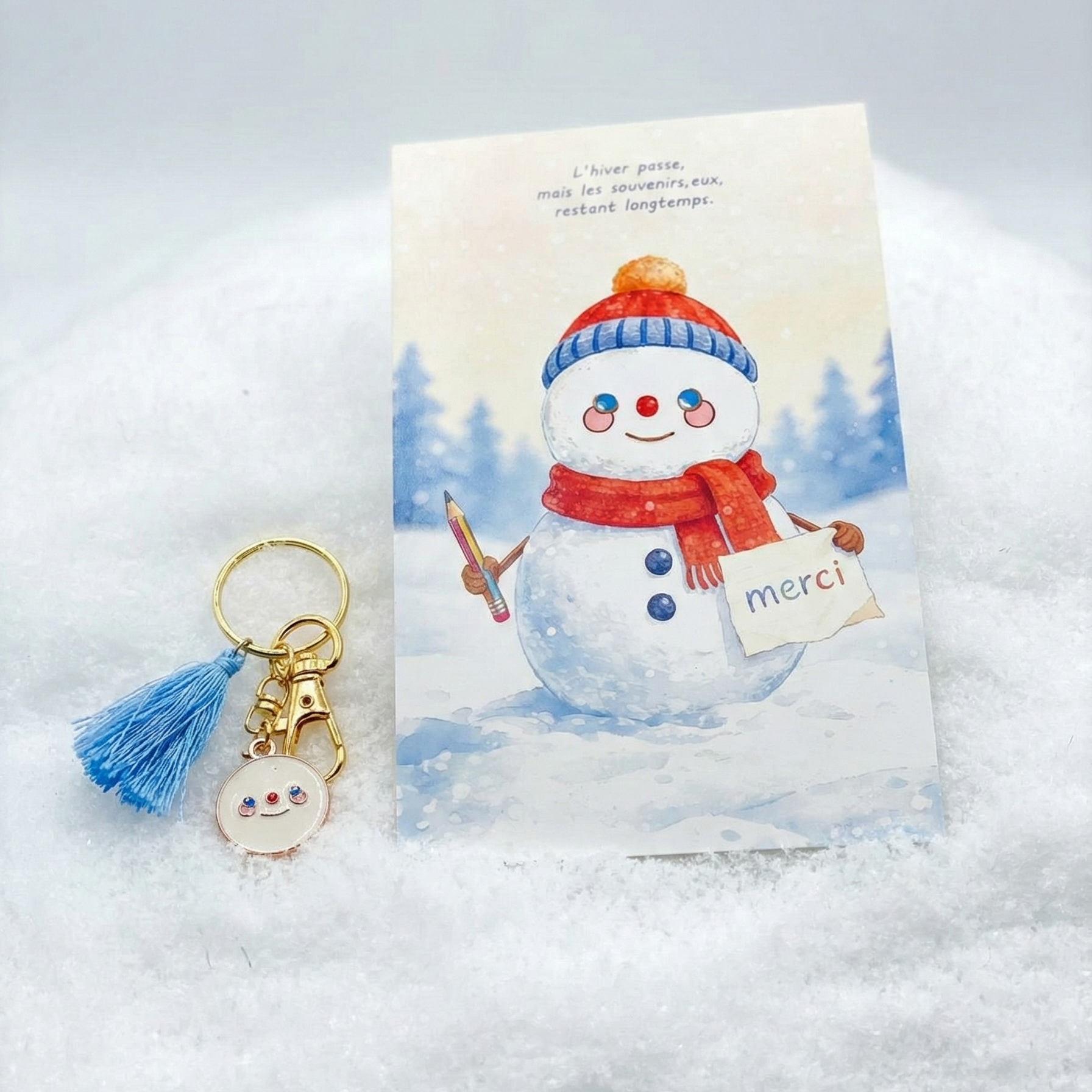 Kit cadeau de deux pièces présenté sur un lit de neige artificielle blanche. À droite, une carte illustrée à l'aquarelle avec un bonhomme de neige portant un bonnet bleu et rouge et une écharpe rouge, tenant un crayon et un panneau "merci". Le texte en haut indique : "L'hiver passe, mais les souvenirs, eux, restent longtemps." À gauche, un porte-clés doré avec un pompon bleu ciel et une petite breloque ronde en émail blanc représentant un visage de bonhomme de neige souriant.