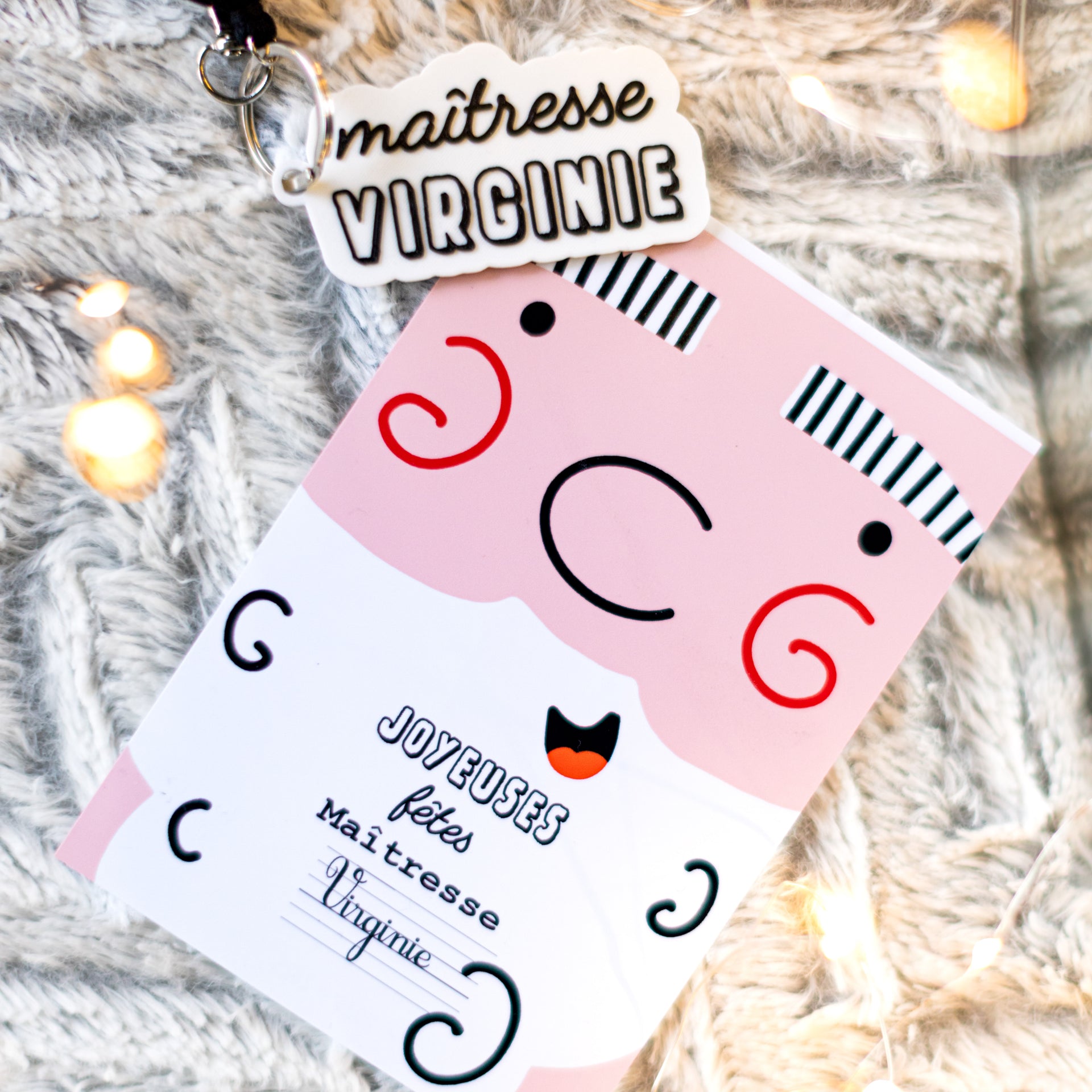 Kit cadeau de Noël pour maîtresse comprenant un porte-clés blanc en impression 3D PLA personnalisé avec le texte 'maîtresse VIRGINIE' en noir, accompagné d'une carte de vœux rose et blanche arborant le message 'JOYEUSES fêtes Maîtresse Virginie'. L'ensemble est mis en scène sur un plaid gris duveteux avec des guirlandes lumineuses festives.