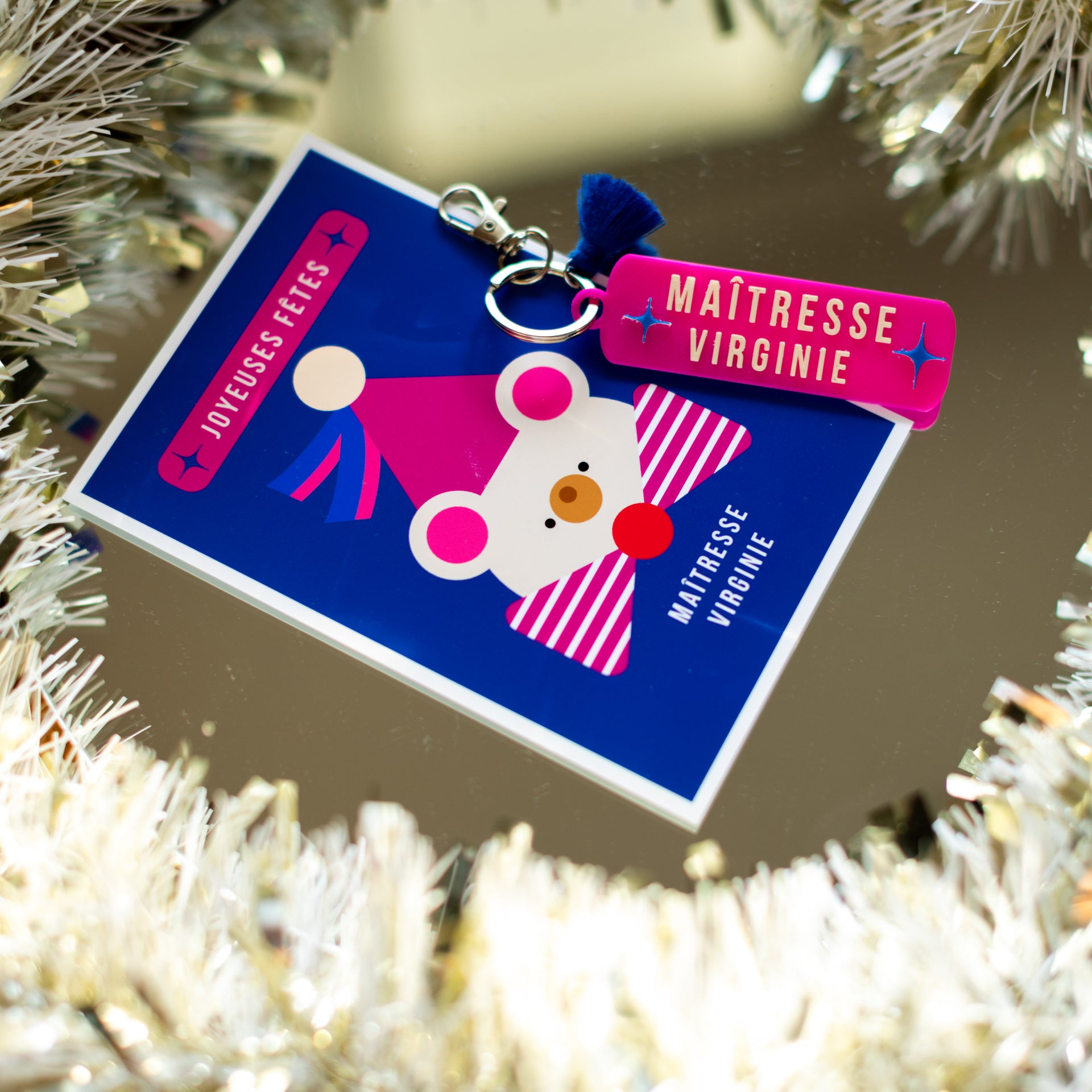 Gros plan d'un kit cadeau de fin d'année pour enseignante, composé d'un porte-clés personnalisé rose fuchsia et "MAÎTRESSE VIRGINIE" en lettres blanches avec un petit pompon bleu marine, et d'une carte de vœux bleu roi assortie avec une illustration géométrique de souris festive portant un chapeau. Le texte sur la carte lit "JOYEUSES FÊTES" et "MAÎTRESSE VIRGINIE". L'ensemble est entouré d'une guirlande argentée festive sur un fond miroir.