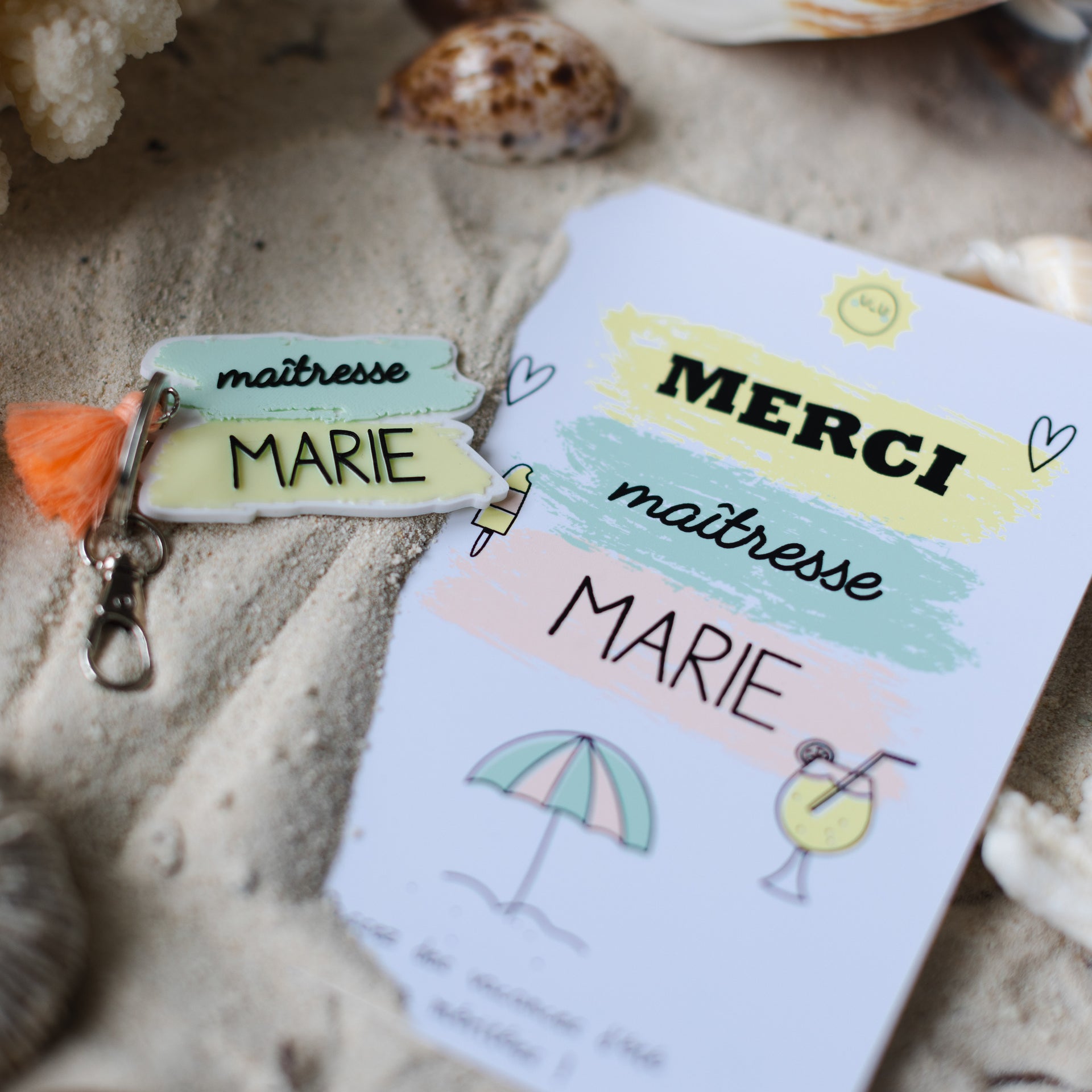 Un kit cadeau pour maîtresse personnalisé pour Marie, posé sur du sable de plage avec des coraux et un coquillage en arrière-plan flou. Il comprend un porte-clés blanc et pastel avec le texte 'maîtresse MARIE' et un pompon orange vif, accompagné d'une carte de remerciement assortie affichant le texte 'MERCI maîtresse MARIE' et des illustrations estivales (parasol, glace, cocktail).