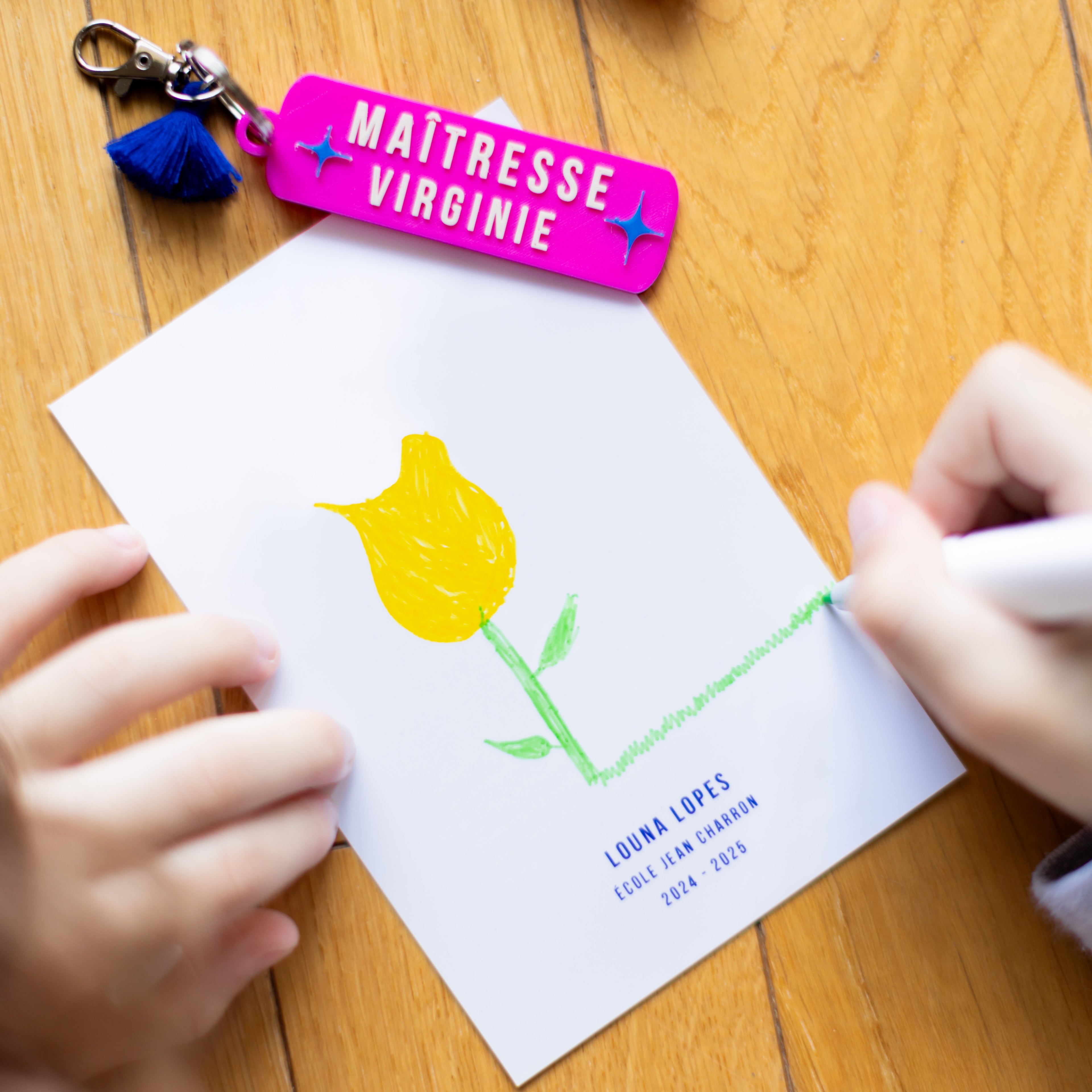 Mains d'un enfant en train de dessiner une tulipe jaune sur une carte de remerciement personnalisée au nom de "Louna Lopes", école Jean Charron 2024-2025. Un porte-clés rose fuchsia personnalisé "Maîtresse Virginie" avec un pompon bleu marine est posé en haut de la carte sur un parquet en bois.
