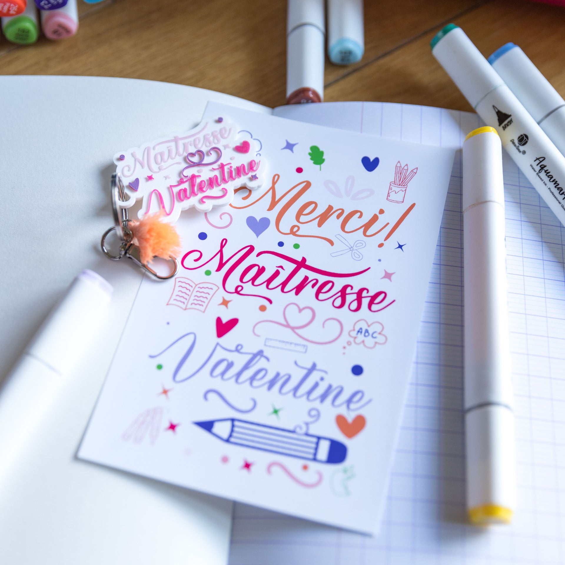 Kit cadeau pour maîtresse comprenant une carte de remerciement colorée "Merci ! Maîtresse Valentine" avec des illustrations d'école (crayon, livre, cœurs) et un porte-clés assorti en impression 3D blanc et rose avec un pompon corail. L'ensemble est mis en scène sur un cahier avec des feutres de dessin.