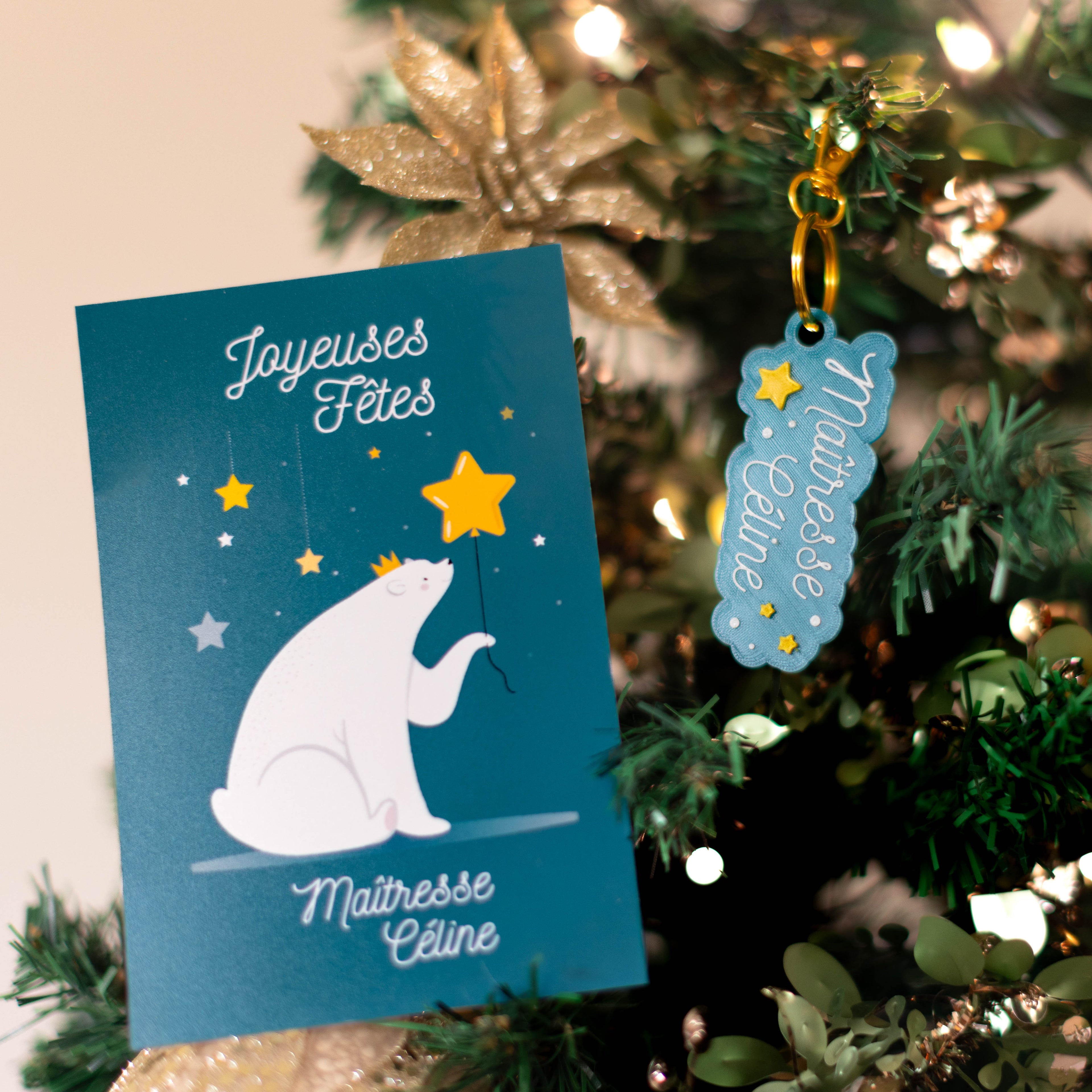 Kit cadeau de Noël personnalisé pour maîtresse Céline : une carte de vœux bleu seychelle foncé avec un ours polaire et une étoile, et un porte-clés bleu personnalisé avec des étoiles, tous deux accrochés à une branche de sapin de Noël décoré.