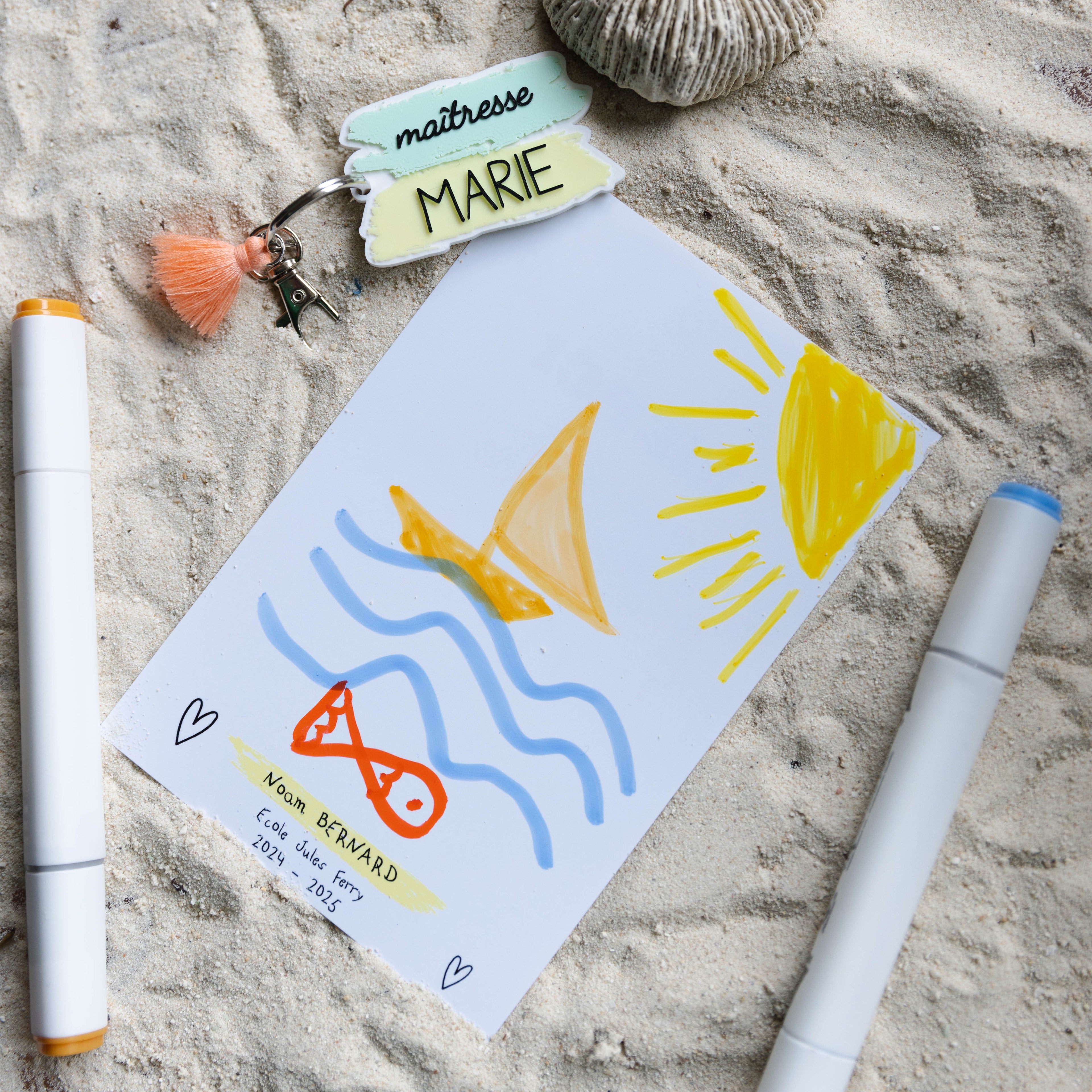 Une photo en vue de dessus, façon flat-lay sur du sable blanc fin, montrant un kit cadeau personnalisé pour l'enseignante "MARIE". Le kit comprend une carte à message dessinée à la main par un enfant nommé Noam BERNARD de l'école Jules Ferry pour l'année scolaire 2024-2025. Le dessin d'enfant représente un voilier sur des vagues bleues et un grand soleil jaune vif, avec un poisson orange. Deux petits cœurs sont également dessinés. Au-dessus de la carte, le porte-clés personnalisé.
