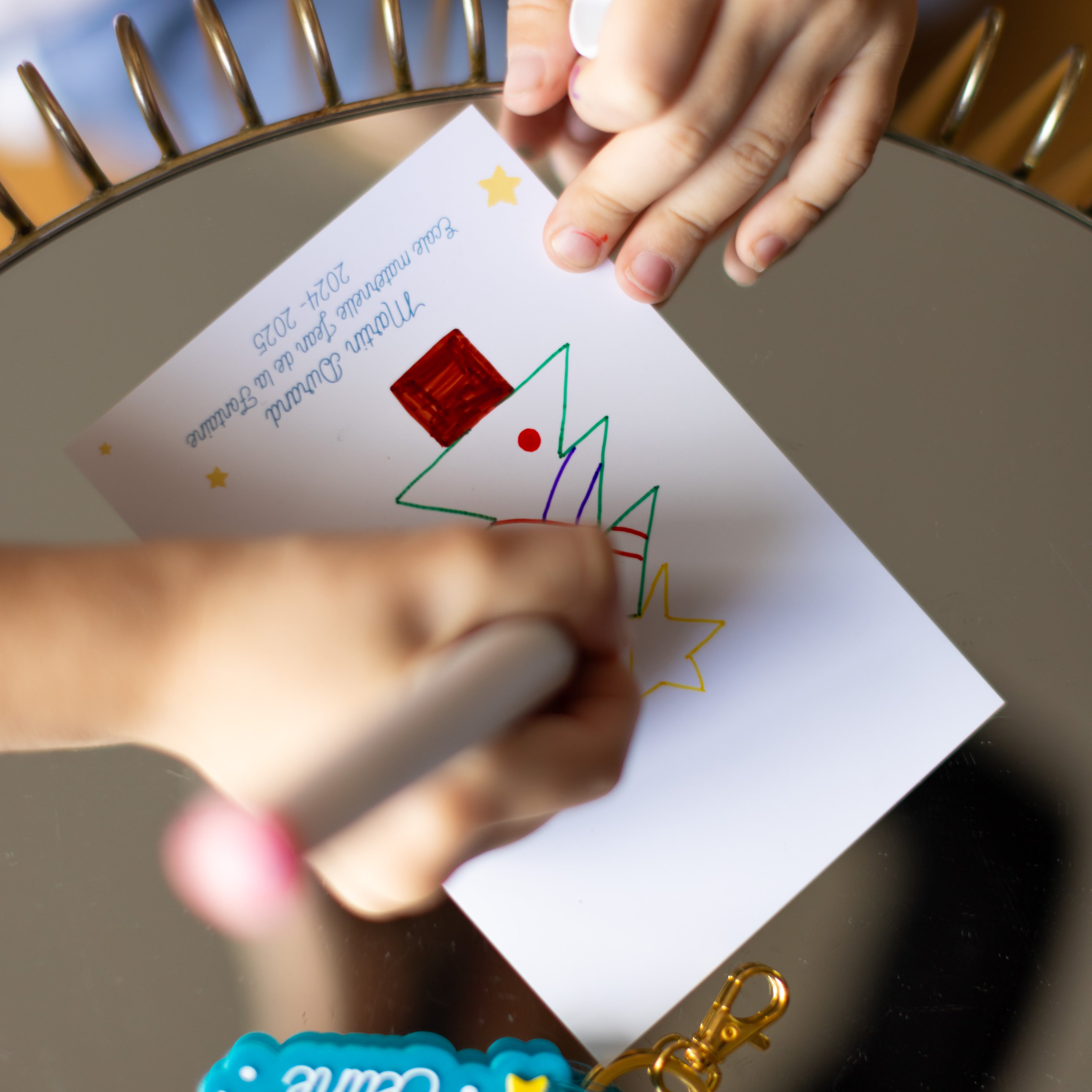Gros plan détaillé sur les mains d'un enfant en train de colorier un dessin d'arbre de Noël stylisé avec un marqueur rose sur une carte personnalisée. La carte, posée sur une surface réfléchissante avec un cadre doré, contient le texte imprimé : "Martin Durand, Ecole maternelle Jean de la Fontaine, 2024 - 2025" orné de petites étoiles jaunes. En bas, la base d'un porte-clés personnalisé avec le texte « Maîtresse » partiellement visible et son mousqueton doré sont visibles.