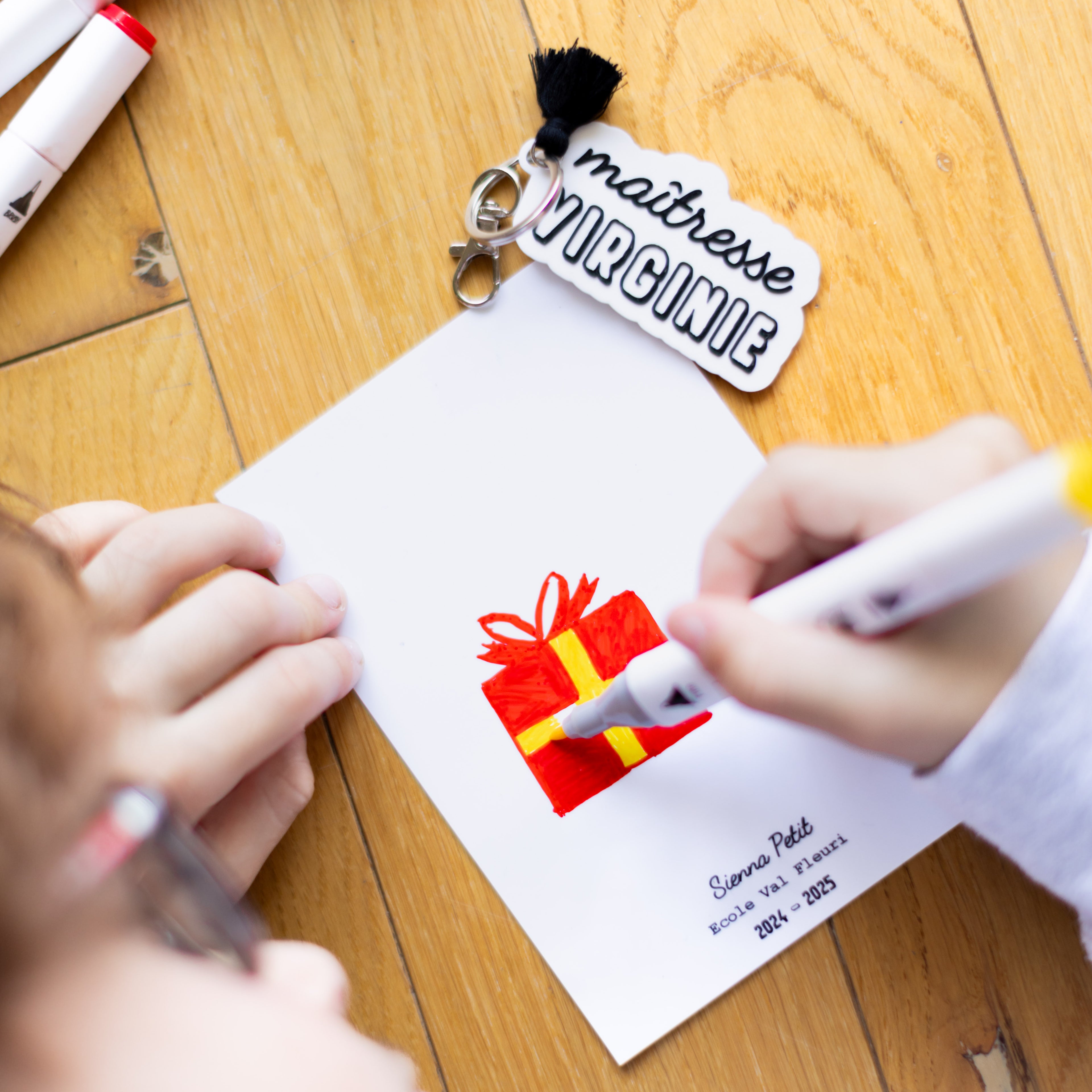 Gros plan d'un enfant en train de dessiner sur une carte de vœux personnalisée pour sa maîtresse. L'enfant utilise un feutre jaune pour colorier le ruban d'un cadeau rouge. La carte est signée « Sienna Petit », « Ecole Val Fleuri », « 2024 - 2025 ». Juste au-dessus, le porte-clés coordonné en relief blanc et noir, avec le texte « maîtresse VIRGINIE » et un pompon noir, est visible. L'ensemble est disposé de manière créative sur un parquet en bois.