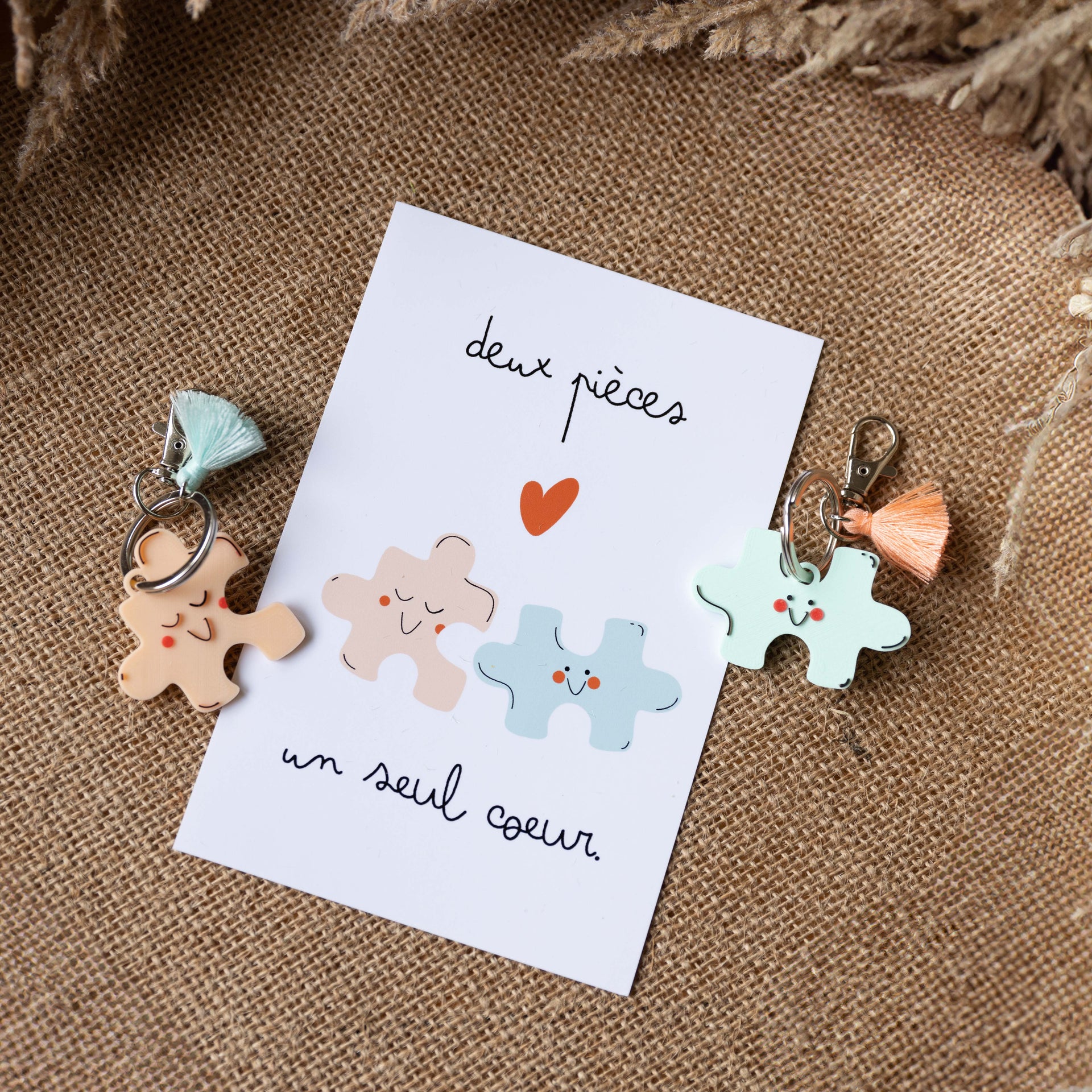 Une photo d'un kit cadeau duo sur fond de toile de jute. Une carte blanche au centre porte le message manuscrit "deux pièces un seul cœur" et une illustration de puzzles mignons qui semblent s'emboîter. À gauche, un porte-clés puzzle rose pêche avec pompon menthe. À droite, un porte-clés puzzle bleu menthe avec pompon corail.