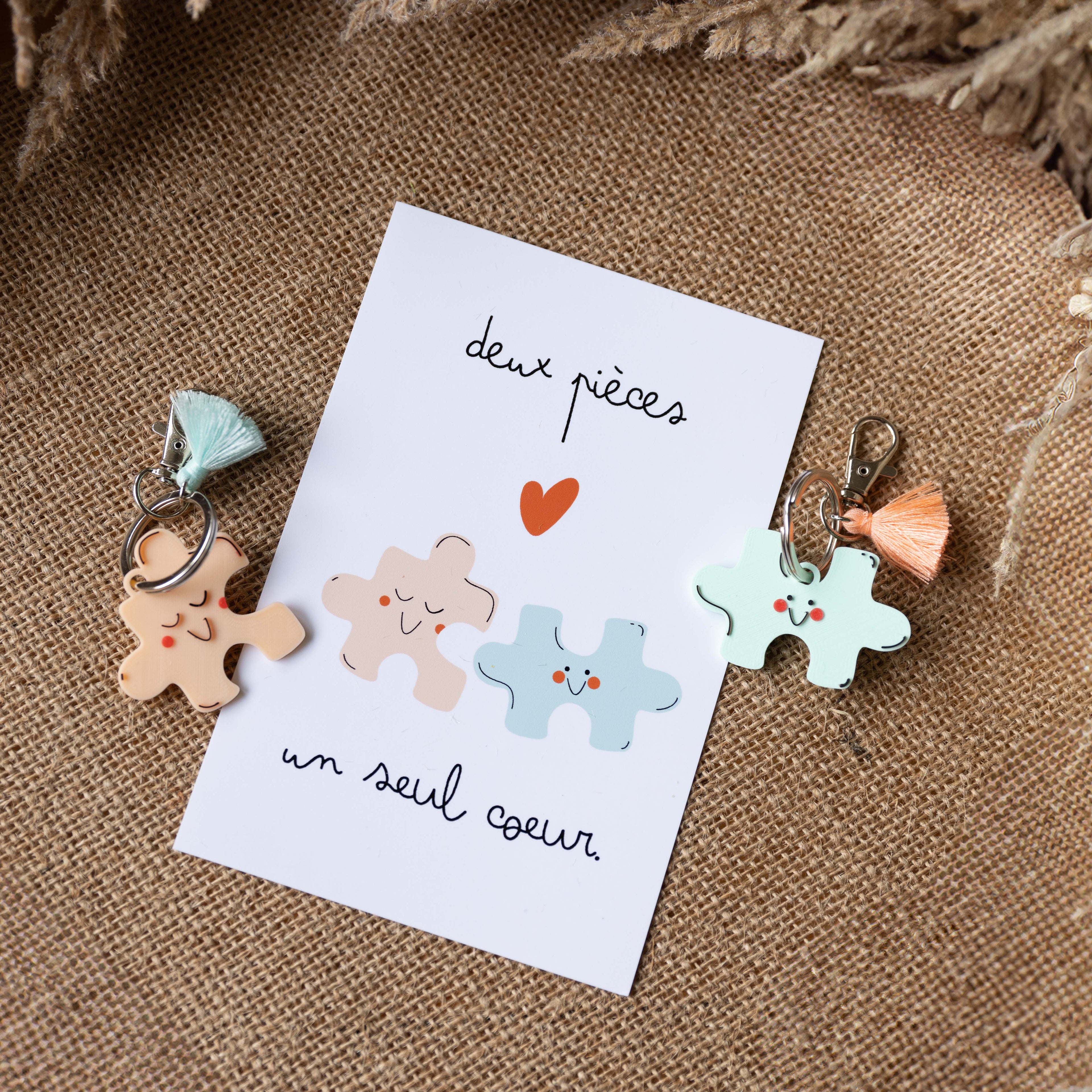 Une photo d'un kit cadeau duo sur fond de toile de jute. Une carte blanche au centre porte le message manuscrit "deux pièces un seul cœur" et une illustration de puzzles mignons qui semblent s'emboîter. À gauche, un porte-clés puzzle rose pêche avec pompon menthe. À droite, un porte-clés puzzle bleu menthe avec pompon corail.