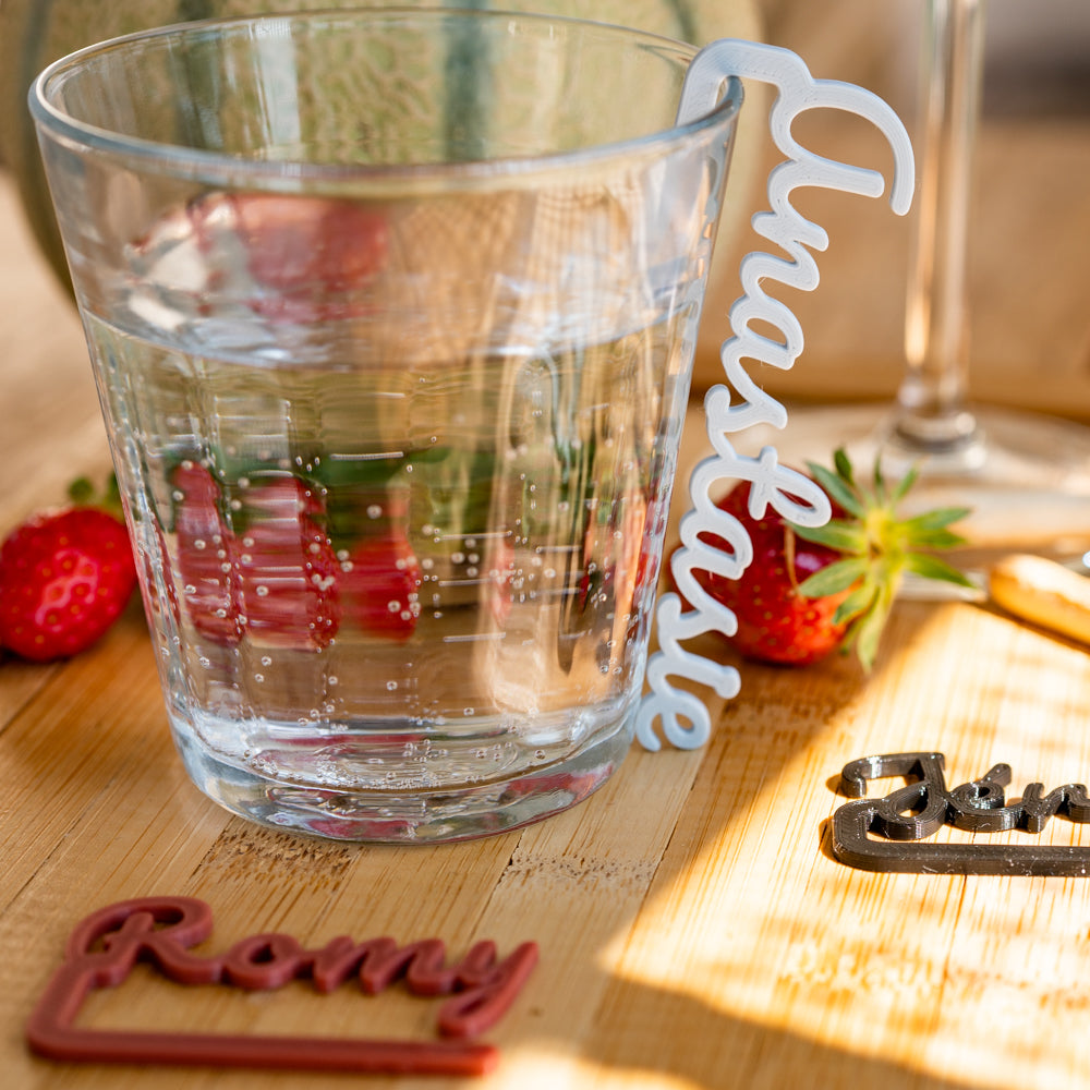 Verre d’eau posé sur une table en bois, orné d’un marque-verre COSTO gris perle portant le prénom “Anastasie” en grandes lettres. À côté, deux fraises et d’autres marque-verres colorés flous en arrière-plan.