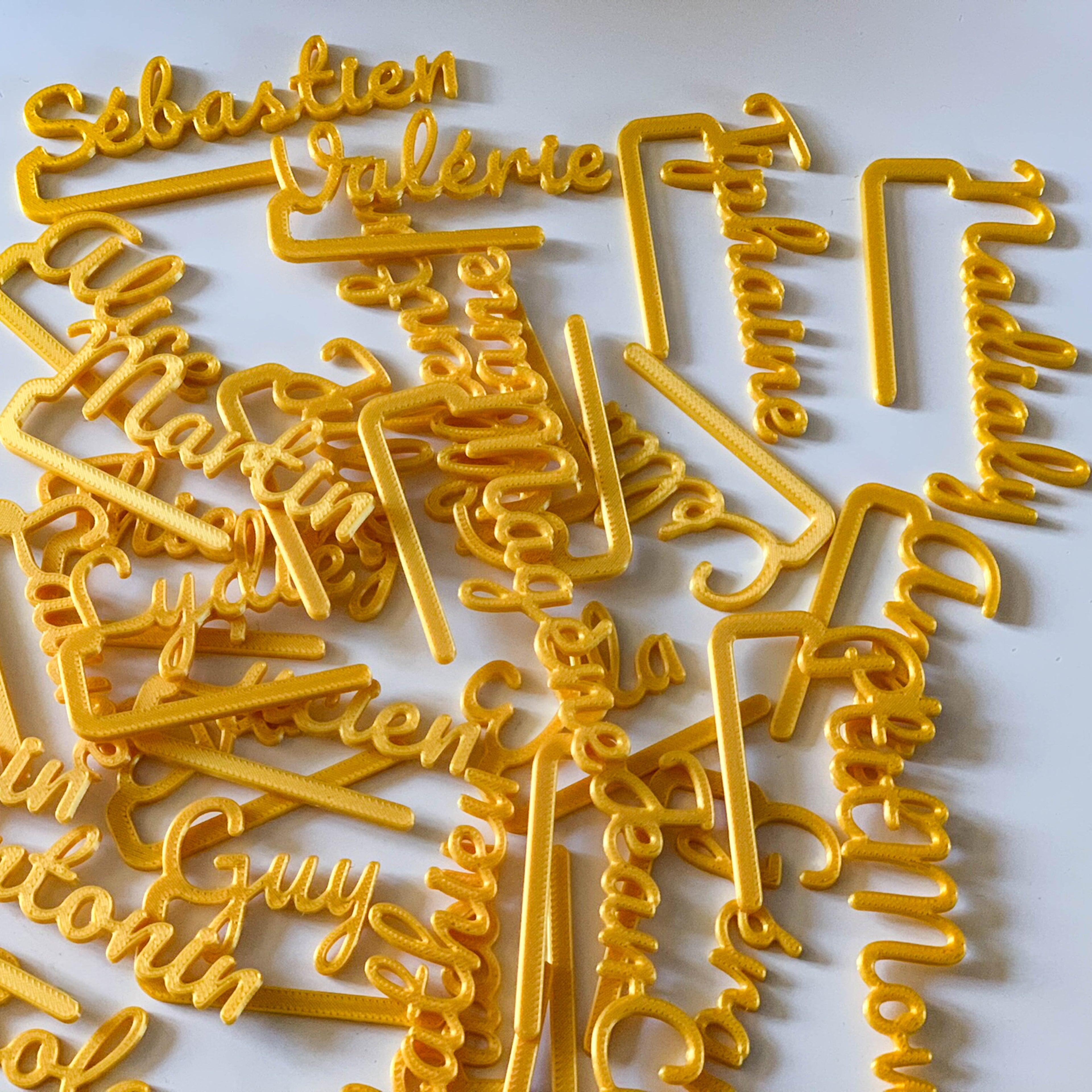 Grand ensemble de marque-verres COSTO en PLA  de couleur doré, étalés en vrac sur une surface blanche. Chaque pièce représente un prénom en écriture cursive prolongée par une barre de clip horizontale. On peut lire des prénoms comme “Sébastien”, “Valérie”, “Guy”, “Quentin”, et d’autres. L’ensemble crée un effet lumineux et festif grâce à la teinte dorée.