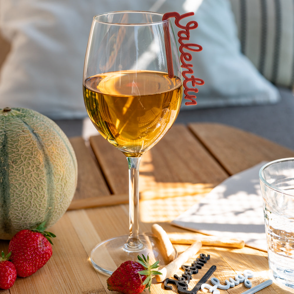 Verre à pied rempli d’un vin blanc ou cidre doré, posé sur une table en bois, avec un marque-verre COSTO rouge portant le prénom “Valentin” accroché au bord du verre. Autour, quelques fraises, un melon entier, des gressins et plusieurs autres marque-verres sont éparpillés. L’ambiance est lumineuse et estivale, évoquant un apéritif en extérieur.