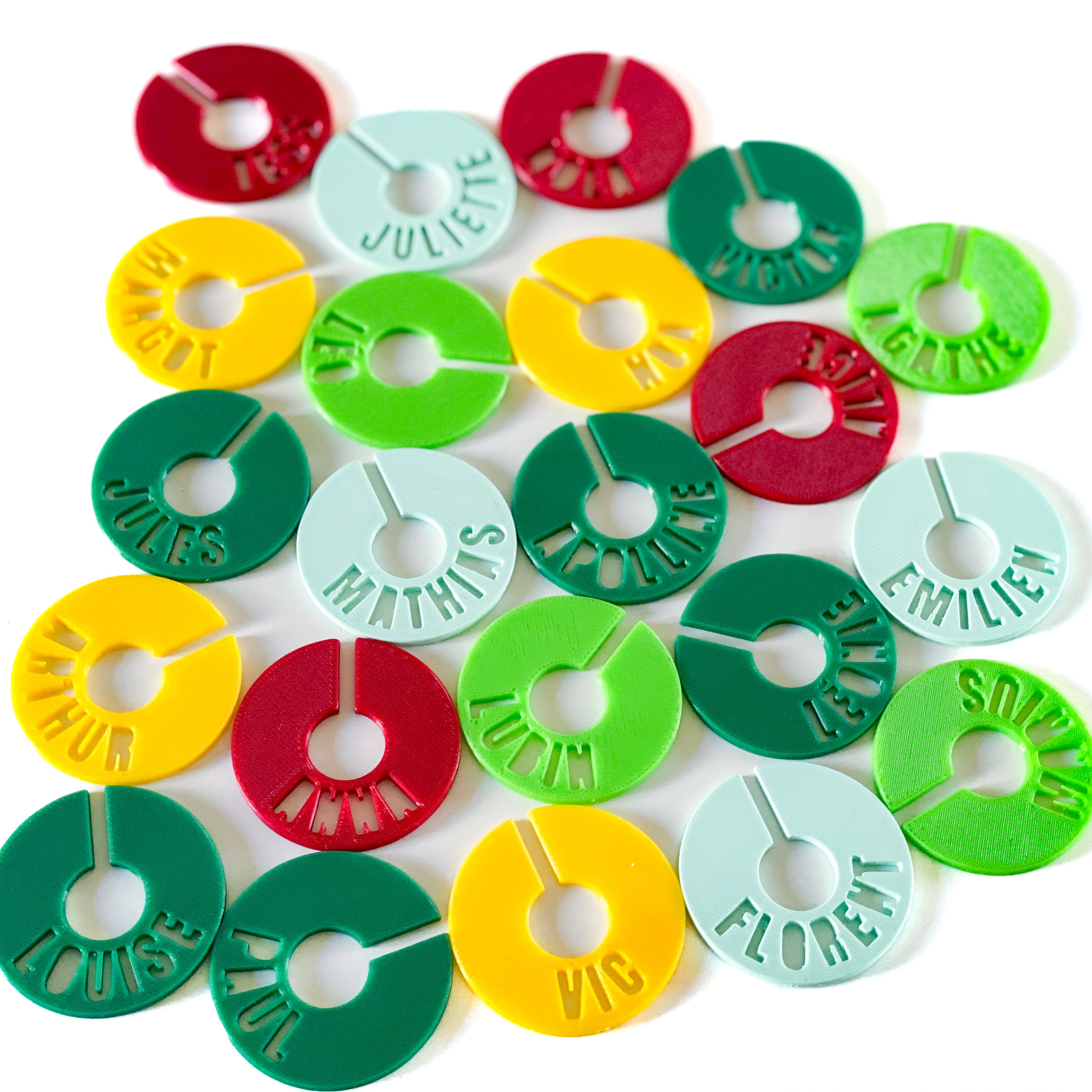 Ensemble de marque-verres creusés multicolores en PLA, disposés en motifs réguliers. Chaque rond est fendu pour s’insérer autour du pied du verre et porte un prénom découpé en arc de cercle.