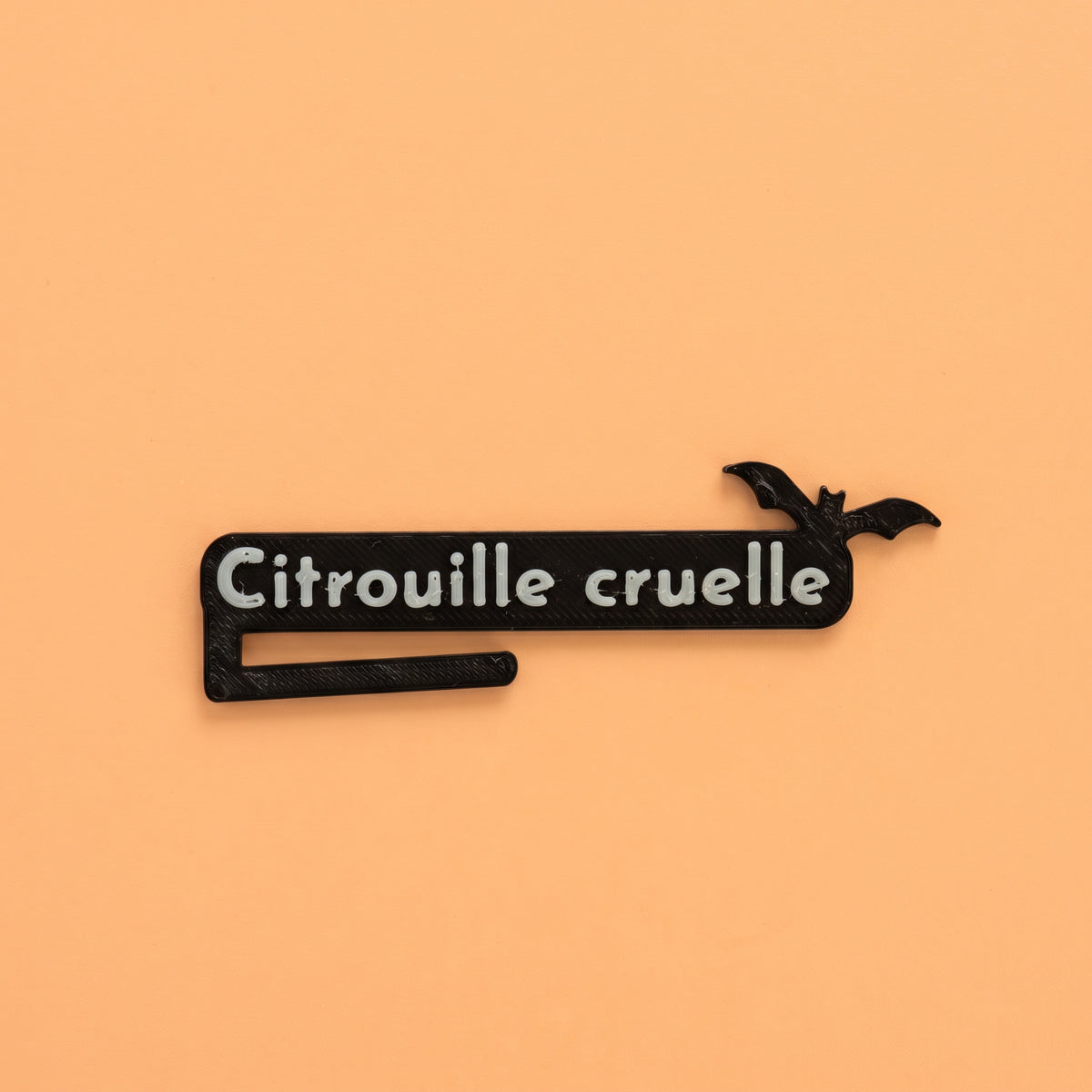 Marque-verre d'Halloween noir imprimé en 3D avec l'inscription « Citrouille cruelle » en relief et une silhouette de chauve-souris, posé à plat sur un fond uni orange pastel lumineux. Accessoire de table original pour identifier son verre lors d'une fête d'Halloween.