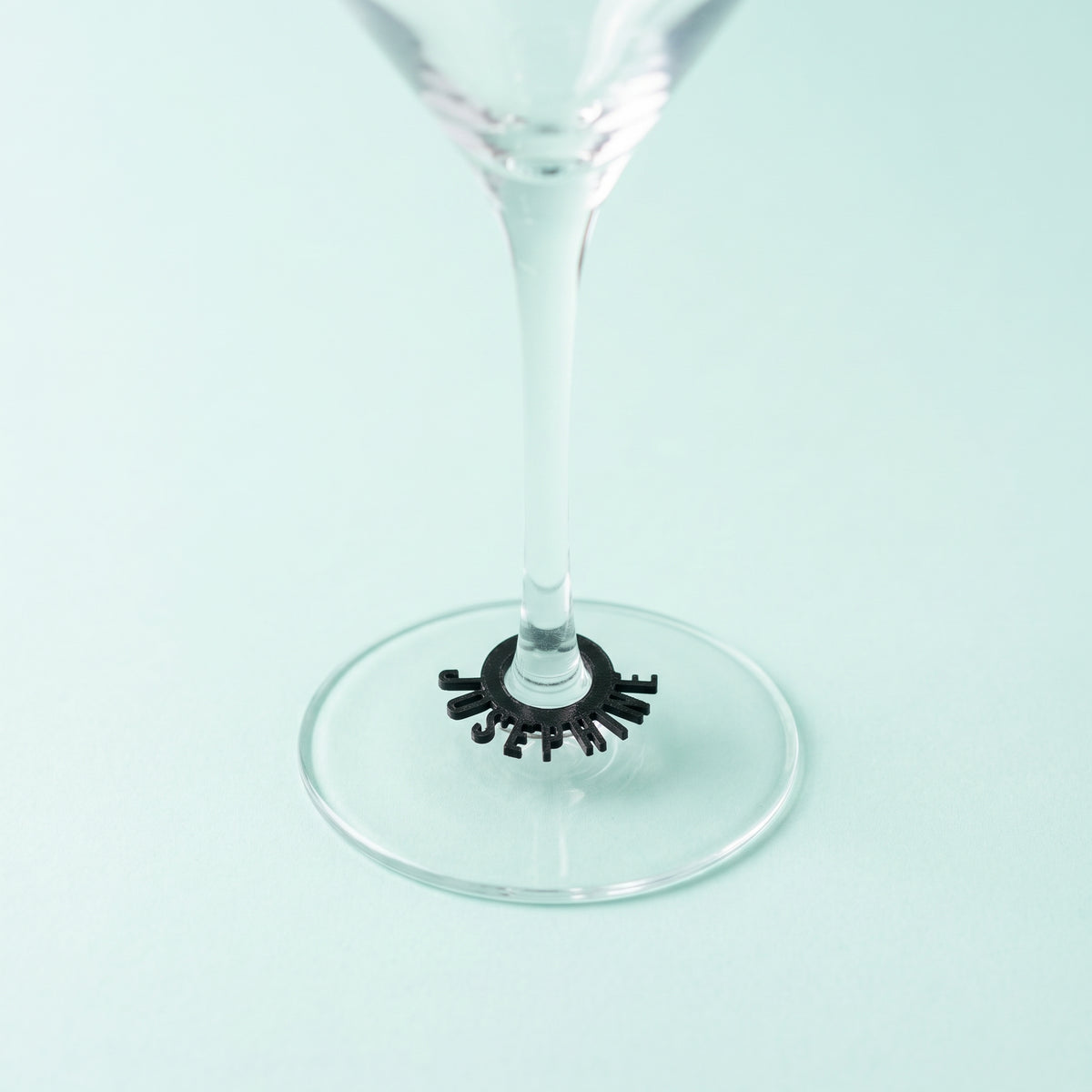 Packshot carré d'un marque-verre personnalisé noir en forme d'anneau avec le prénom JOSEPHINE, fixé à la base d'un verre à pied transparent, posé sur un fond uni vert pastel très lumineux et épuré. En savoir plus sur nos marque-verres originaux pour événements.