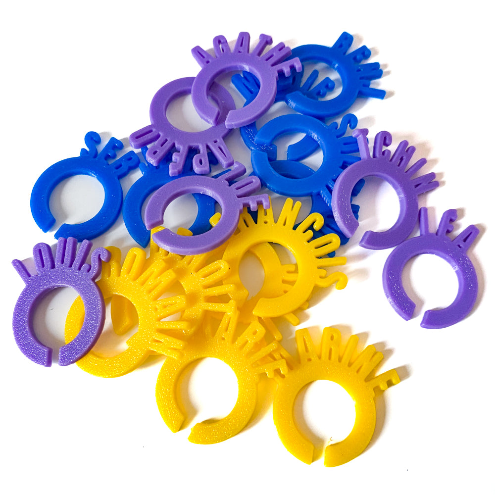 Ensemble de marque-verres en forme d’anneaux personnalisés avec des prénoms, en PLA violet, bleu et jaune. Les lettres verticales dépassent du cercle et les pièces sont disposées en tas sur fond blanc.