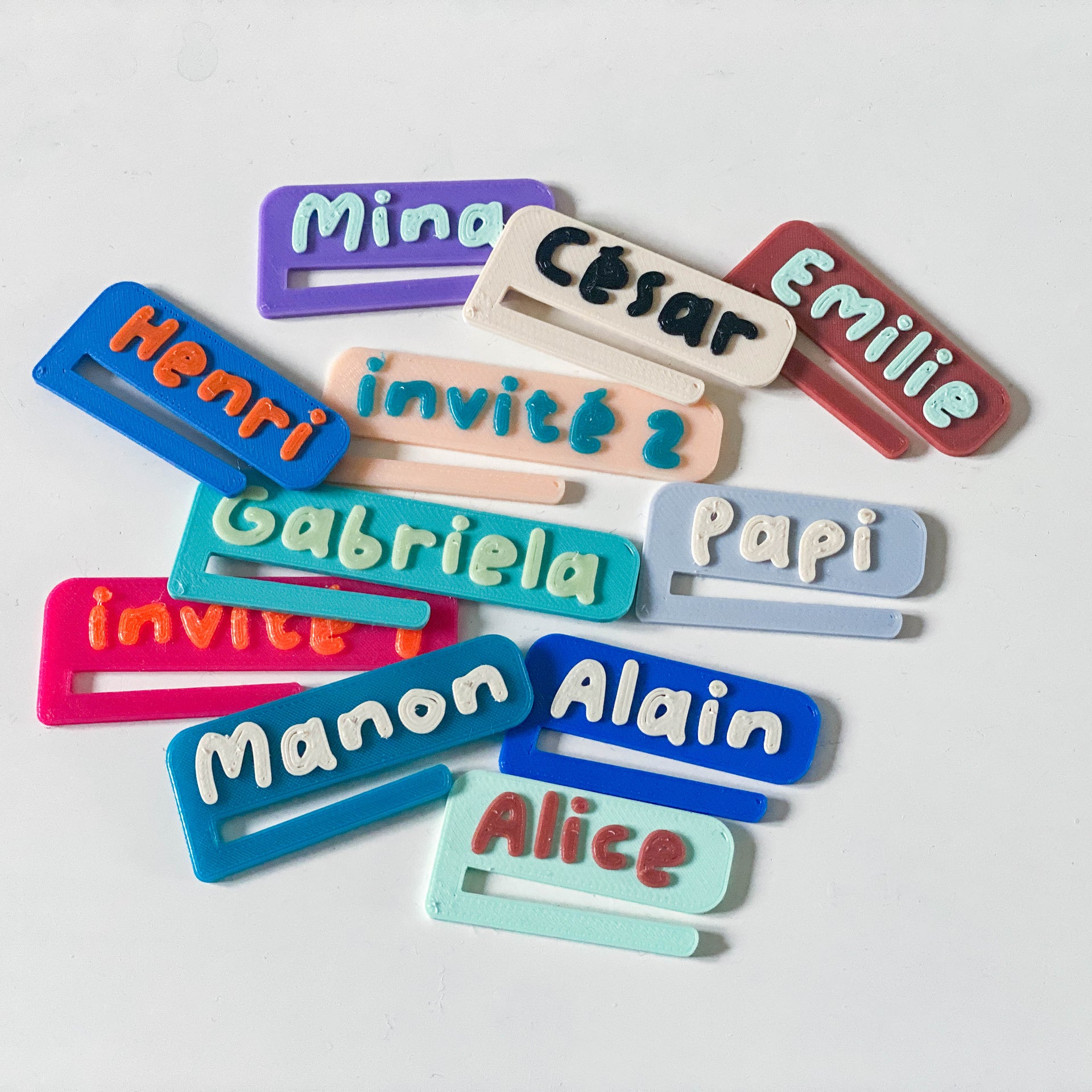 Ensemble de marque-verres bicolores personnalisés en PLA, avec plusieurs prénoms colorés en relief tels que Mina, César, Emilie, Gabriela, Manon, Alain ou Alice, disposés en vrac sur une surface blanche