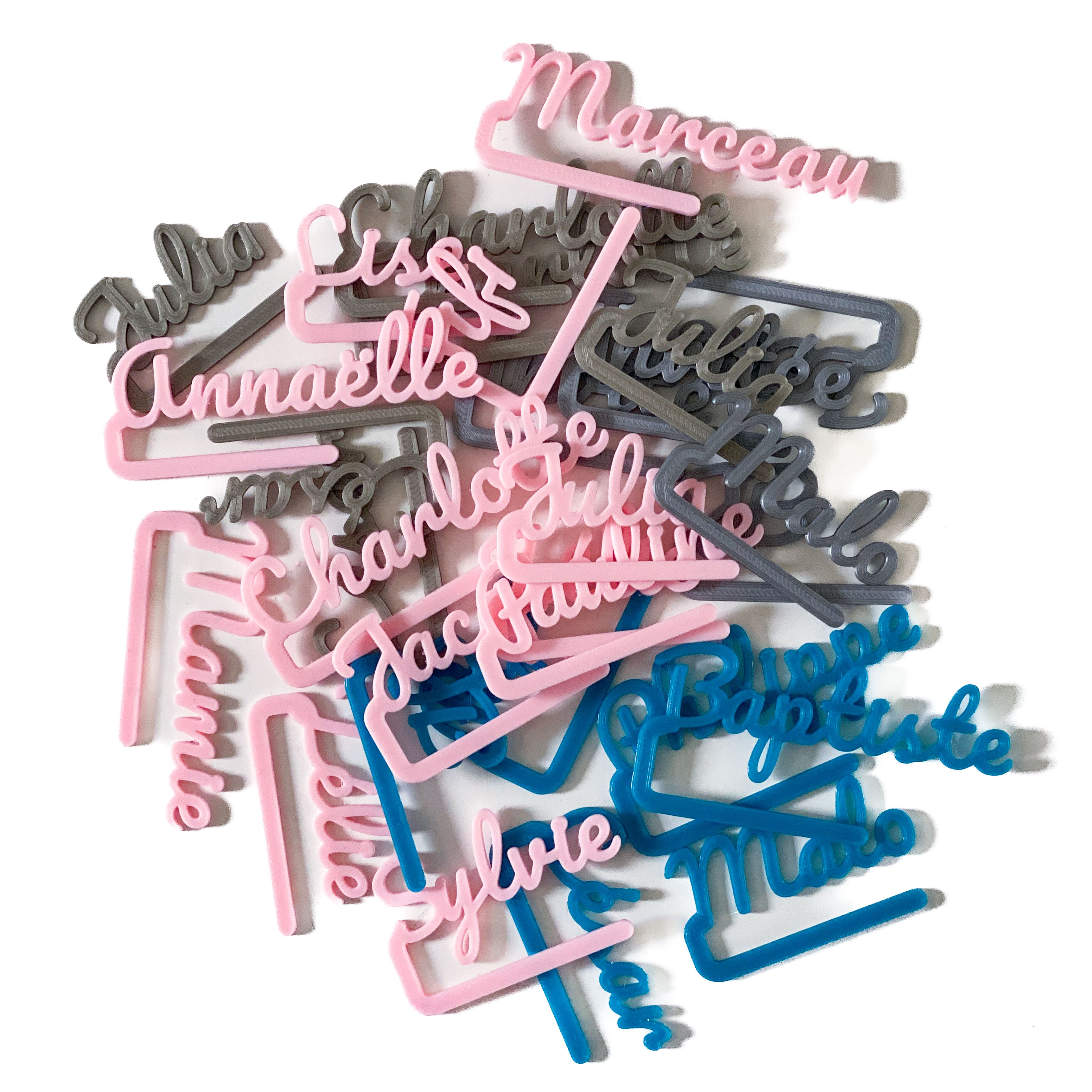 Ensemble de marque-verres COSTO personnalisés, dispersés en pile sur une surface blanche. Les prénoms, imprimés en lettres cursives, apparaissent en rose pâle, gris argent et bleu. Chaque pièce a une longue barre de clip destinée à se fixer sur le bord d’un verre.