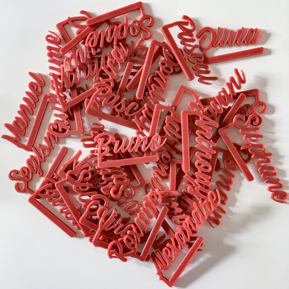 Ensemble de nombreux marque-verres personnalisés en PLA couleur tomette disposés en pile sur une surface blanche. Chaque pièce est en écriture cursive avec un long crochet droit permettant de se clipser au bord d’un verre. Les prénoms variés s’entrecroisent, formant un tas dense où l’on distingue par exemple “Bruno”, “Pascal”, “Julia” ou “Sophie”