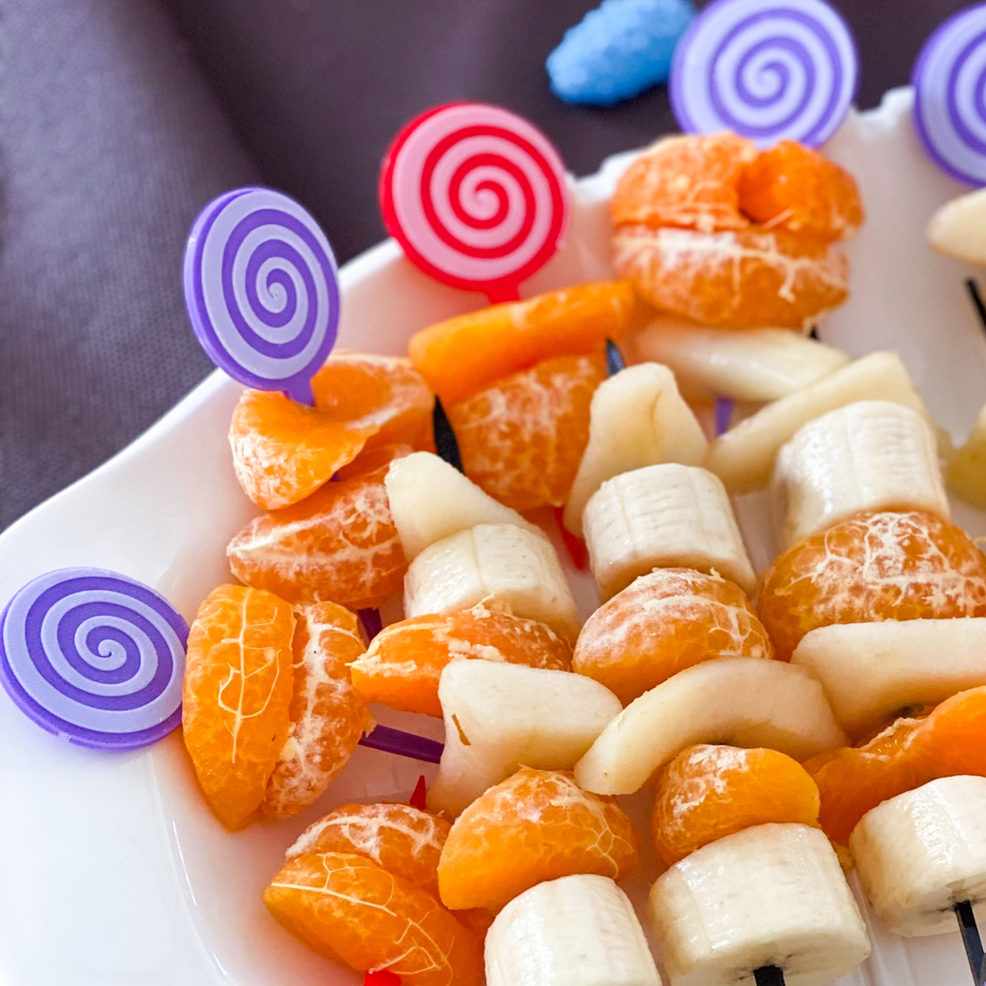 Vue rapprochée d'une assiette blanche garnie de mini-brochettes de fruits frais, surmontées de piques décoratives en forme de sucettes spirales rouges et violettes. Une idée de présentation ludique pour un goûter d'enfant ou une table thématique.