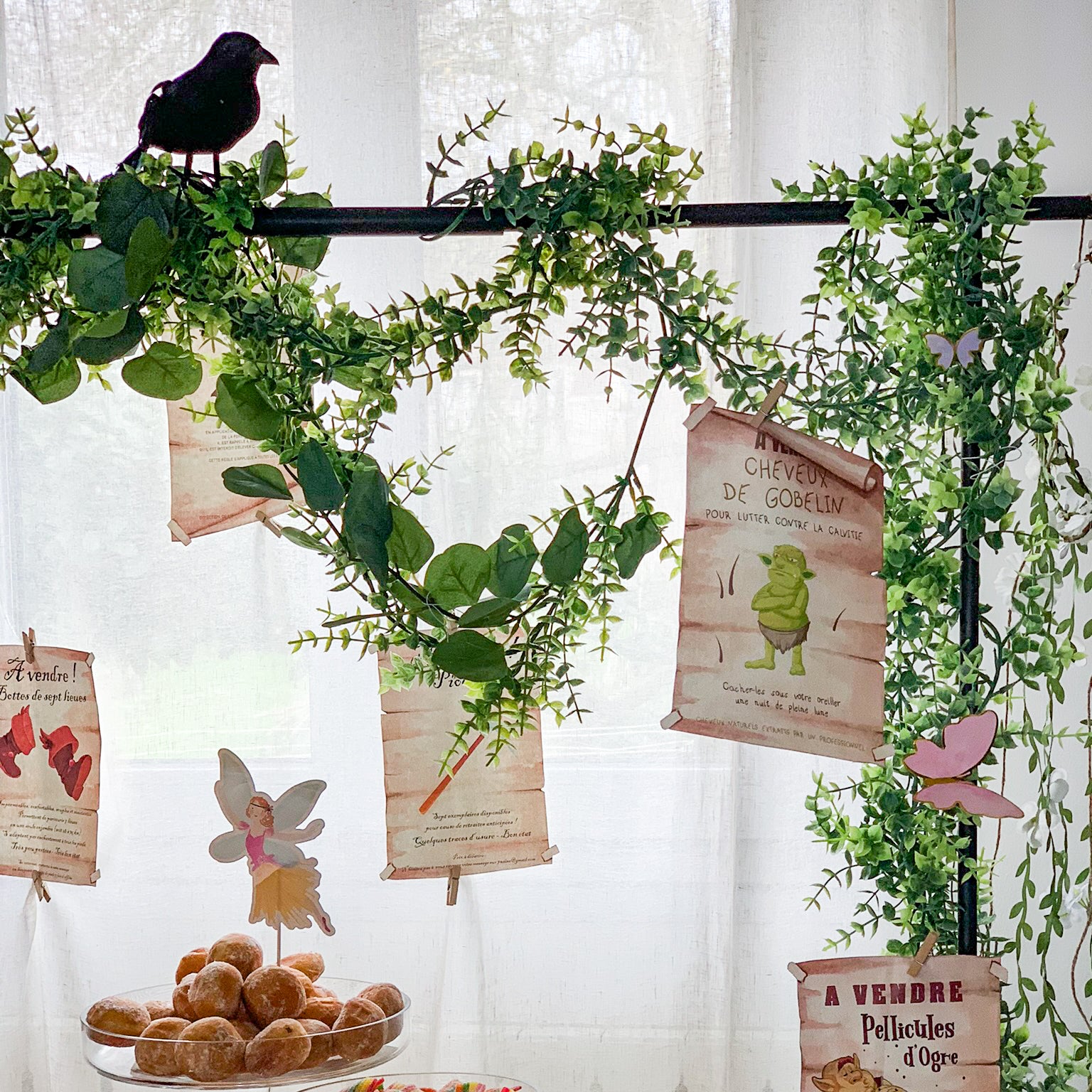 Décoration de fête forêt enchantée avec fausses affiches suspendues dans des feuillages, ambiance féerique avec petits gâteaux, fée et parchemins imprimés – thème magique et nature.