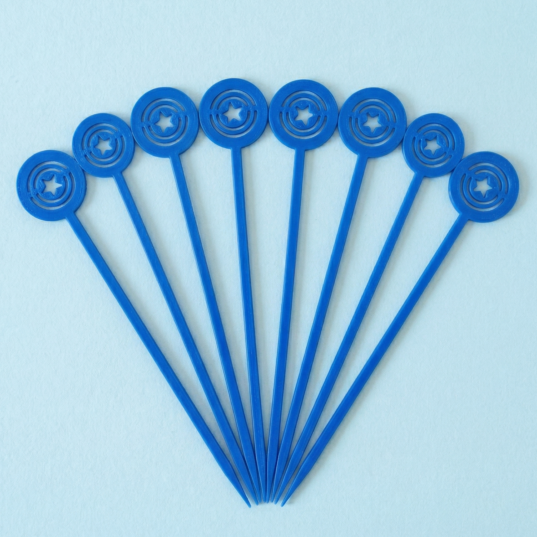 Lot de 8 piques aperitif bleues en forme de bouclier de Captain America disposees en eventail sur un fond bleu clair.