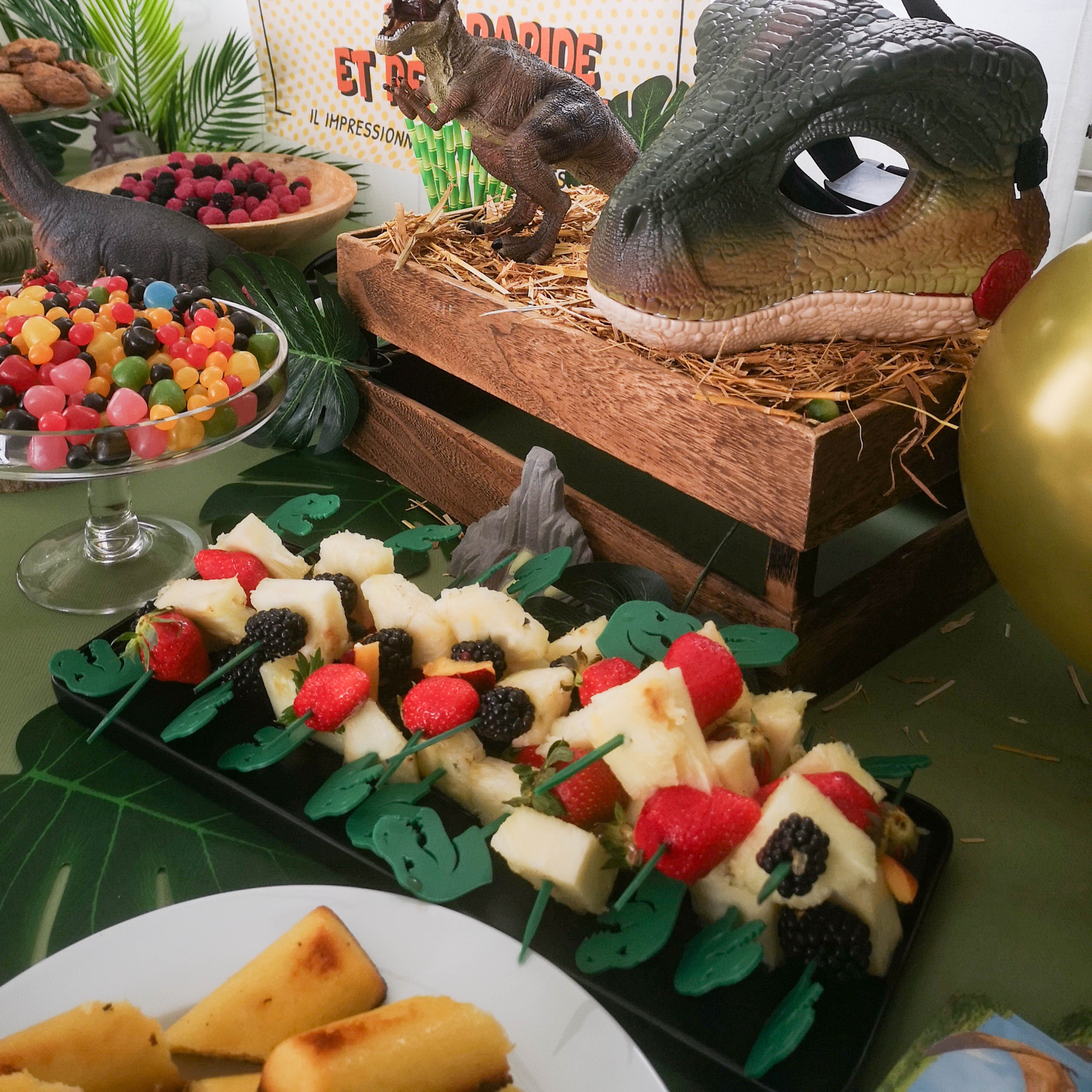 Plateau de brochettes de fruits (ananas, fraises, mûres) décoré de piques T-Rex verts en relief 3D. Mise en scène de buffet pour un anniversaire thème dinosaure avec décoration tropicale et accessoires rustiques.