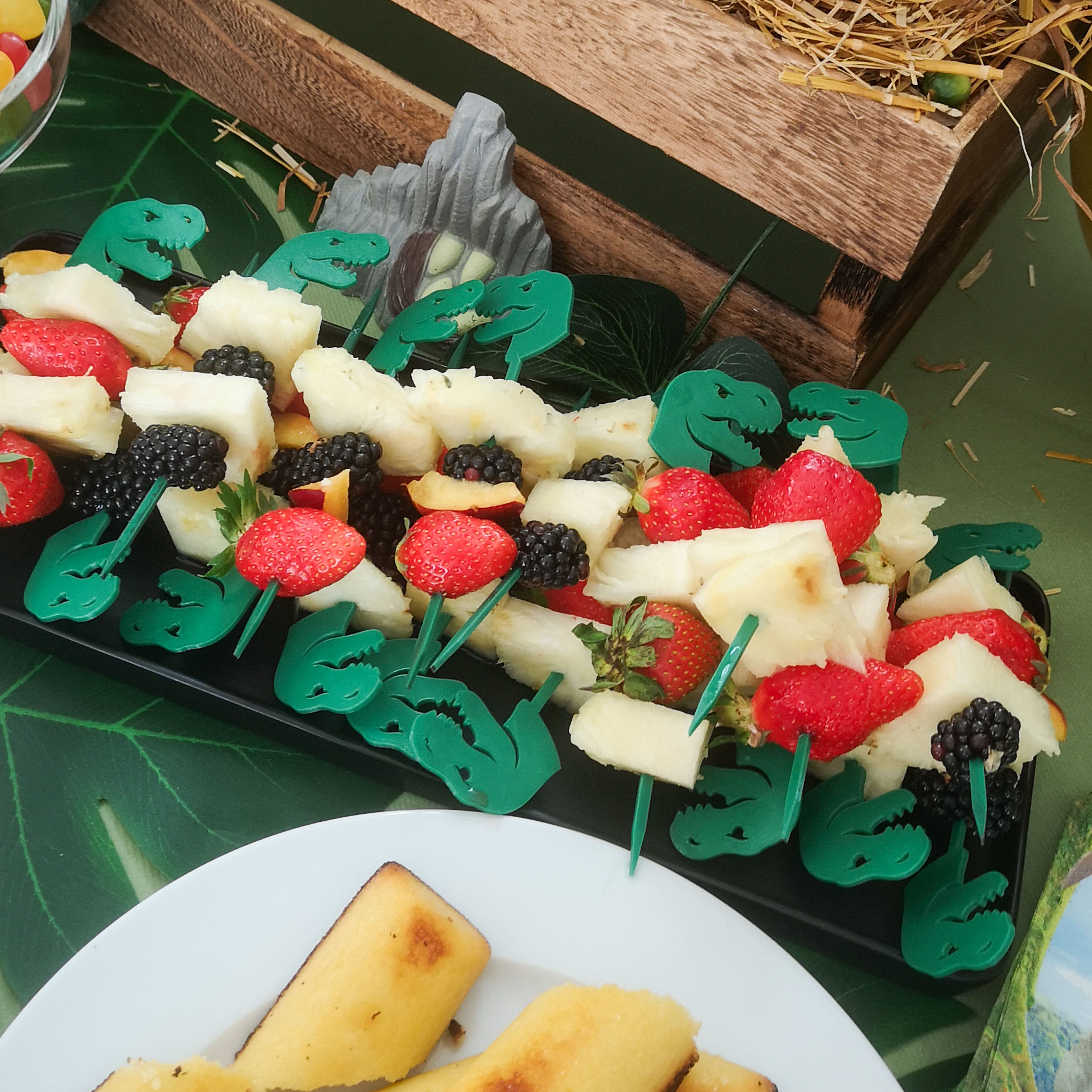 Plateau de brochettes de fruits frais composé d'ananas, de fraises et de mûres, décoré avec des piques dinosaures T-Rex en relief vert sapin. La présentation est disposée sur un buffet à thème avec une nappe en forme de feuille de palmier, une caisse en bois rustique et de la paille en arrière-plan.