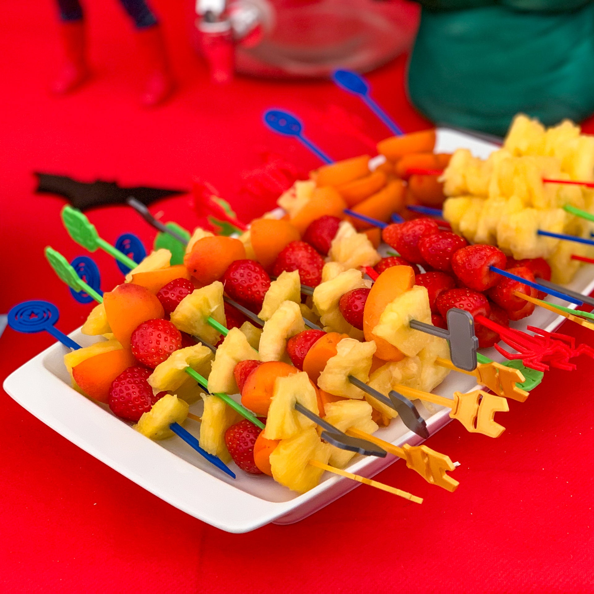 Plateau de brochettes de fruits frais (fraises, ananas, abricots) avec des piques super-heros Avengers (Spider-Man, Iron Man, Captain America, Hulk, Thor) en impression 3D sur un buffet d'anniversaire thématique.