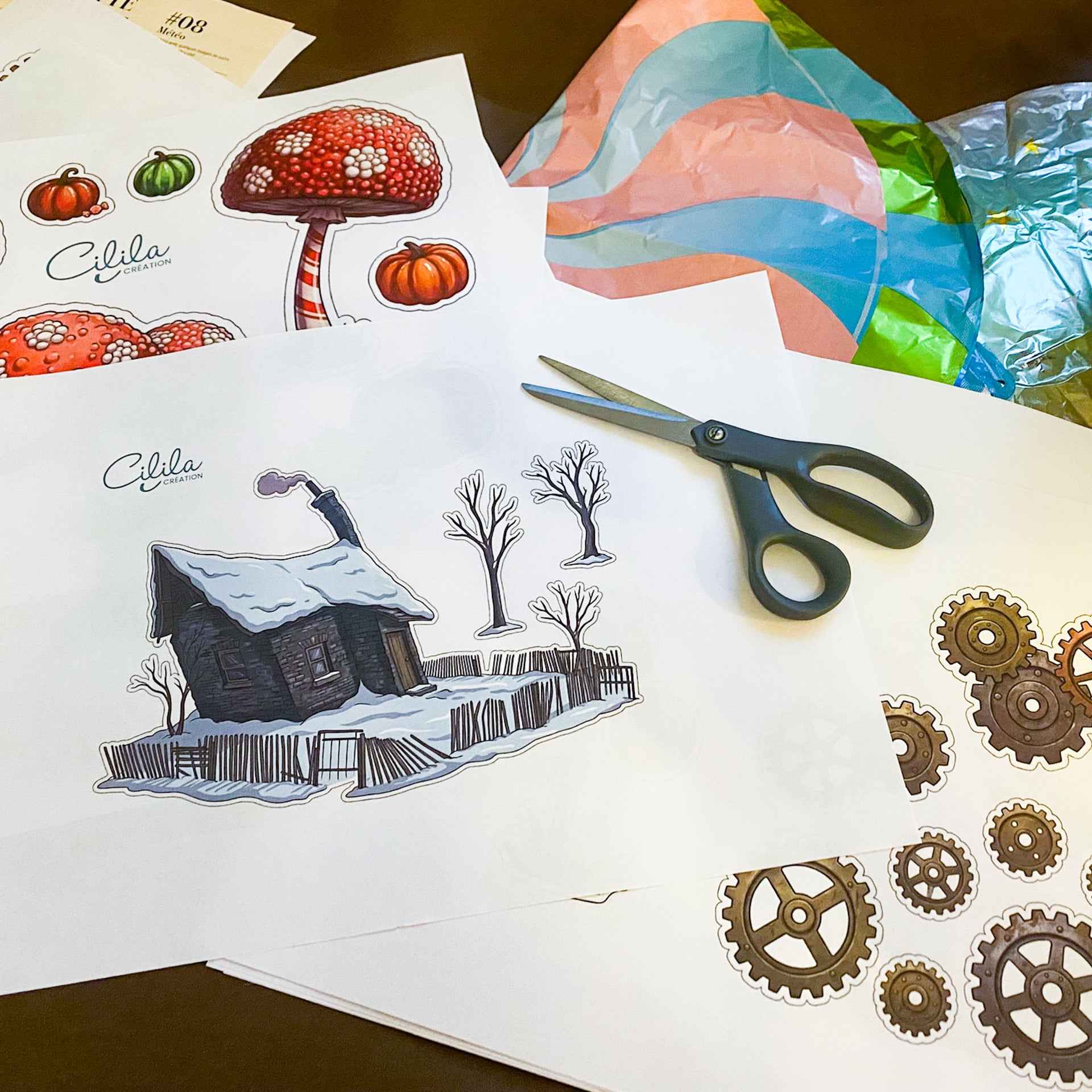 Vue de dessus d'un plan de travail créatif montrant plusieurs planches d'illustrations imprimées (une maison sous la neige, des champignons rouges, des citrouilles, des engrenages), une paire de ciseaux à anneaux noirs et du papier cadeau coloré en arrière-plan.