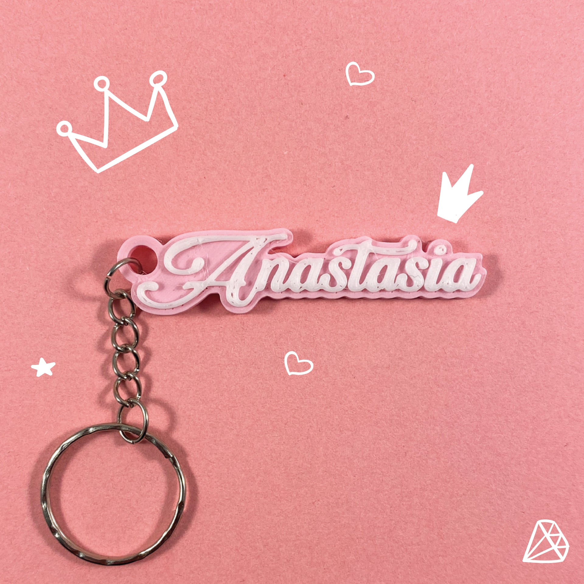 Porte-clés personnalisable avec le prénom "Anastasia" en calligraphie blanche sur une base rose pastel, sur fond rose uni orné de dessins de couronnes de princesse, de cœurs et d'un diamant.