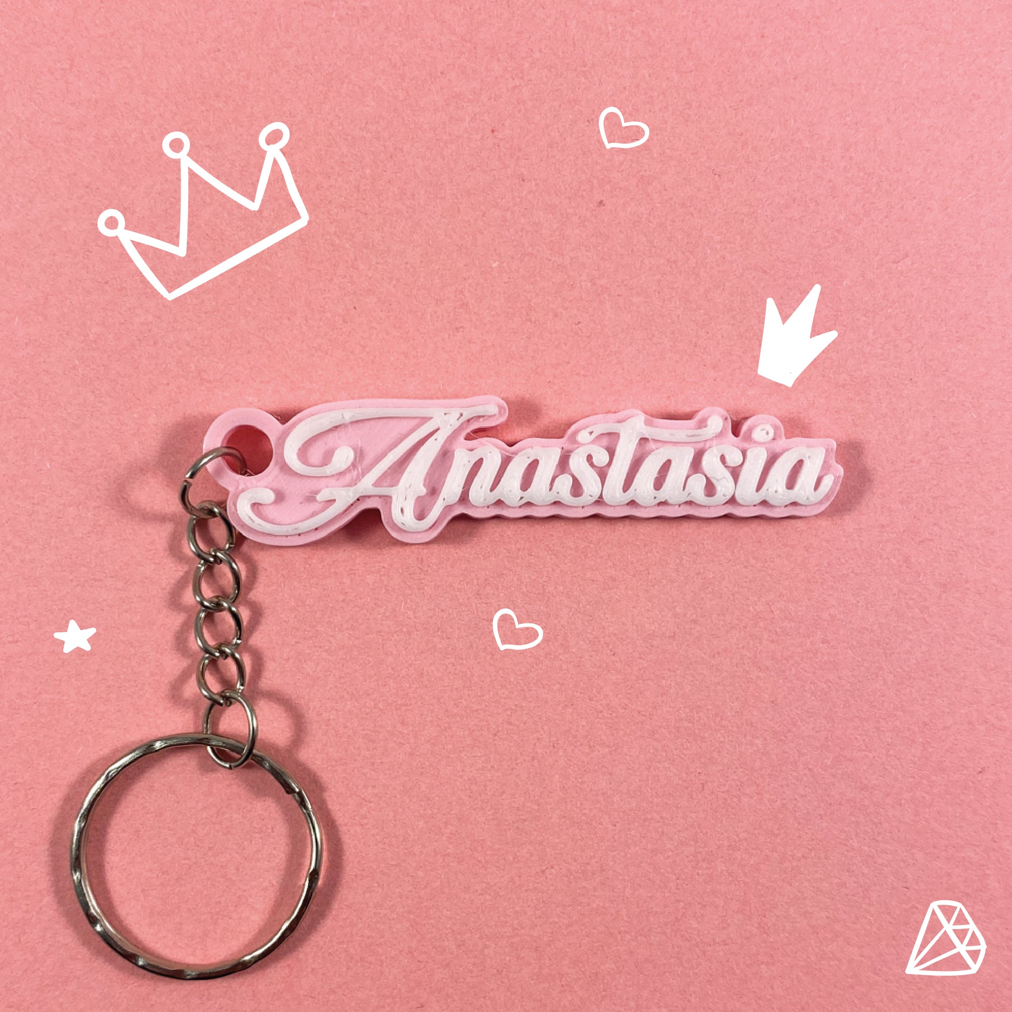 Porte-clés personnalisable avec le prénom "Anastasia" en calligraphie blanche sur une base rose pastel, sur fond rose uni orné de dessins de couronnes de princesse, de cœurs et d'un diamant.